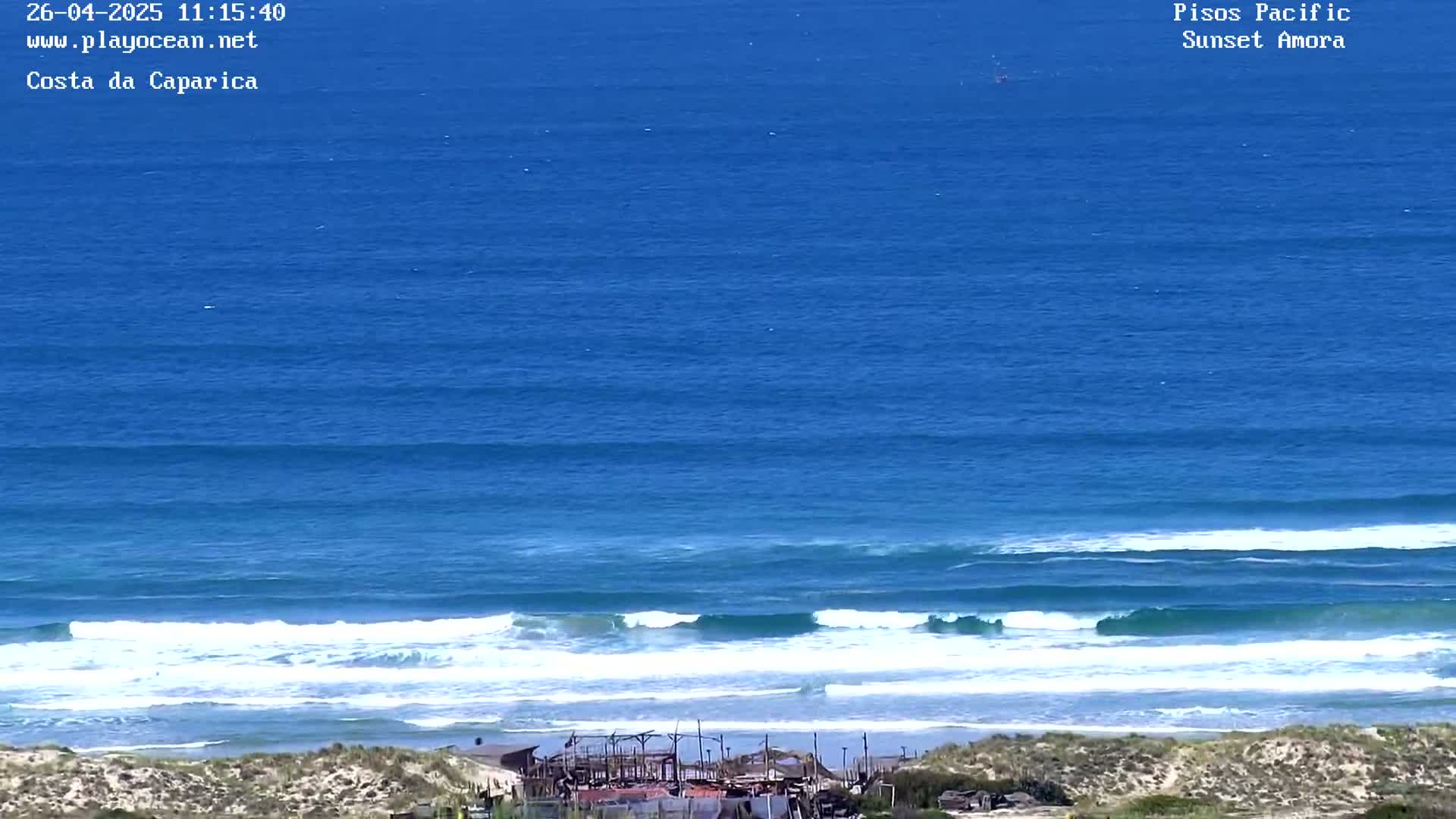 A wide shot of an ocean beach on a sunny day shows a sandy shore with small structures and waves rolling into the shore from a deep blue ocean.