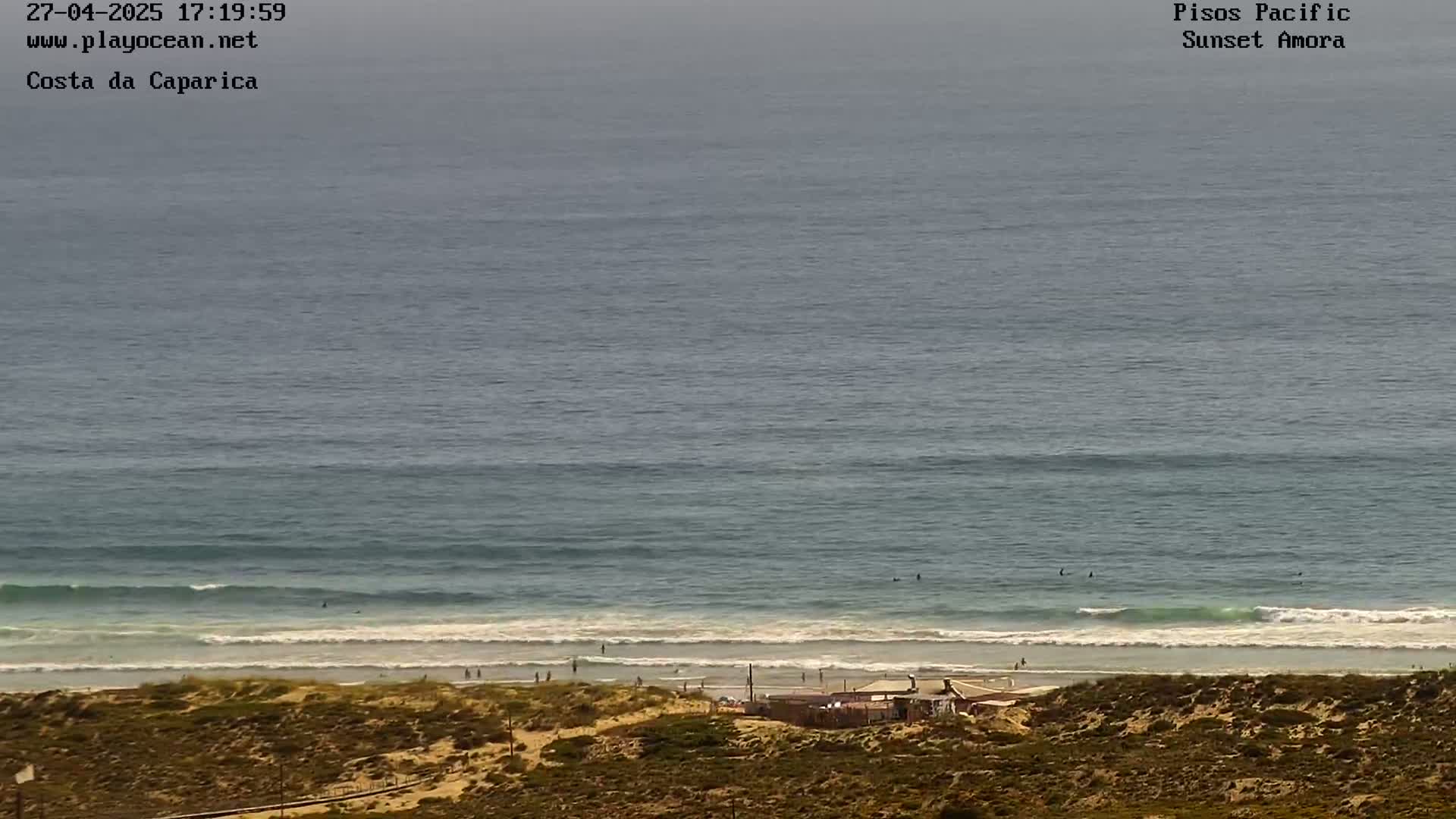 An ocean beach with a few surfers and a small building on the shore is visible under a hazy sky.
