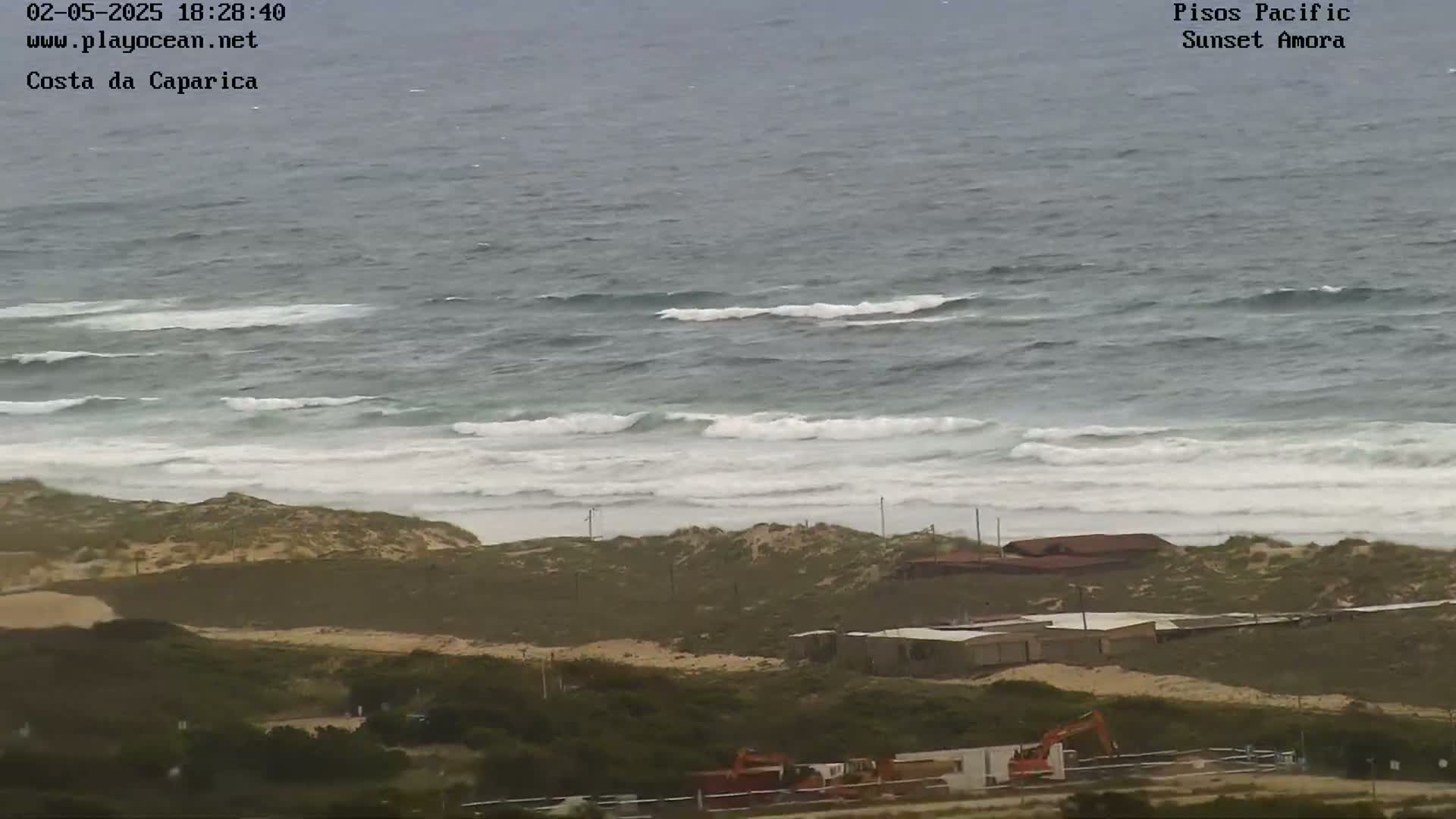 A coastal scene shows moderate waves rolling onto a sandy beach, behind which are low-lying buildings and construction equipment.