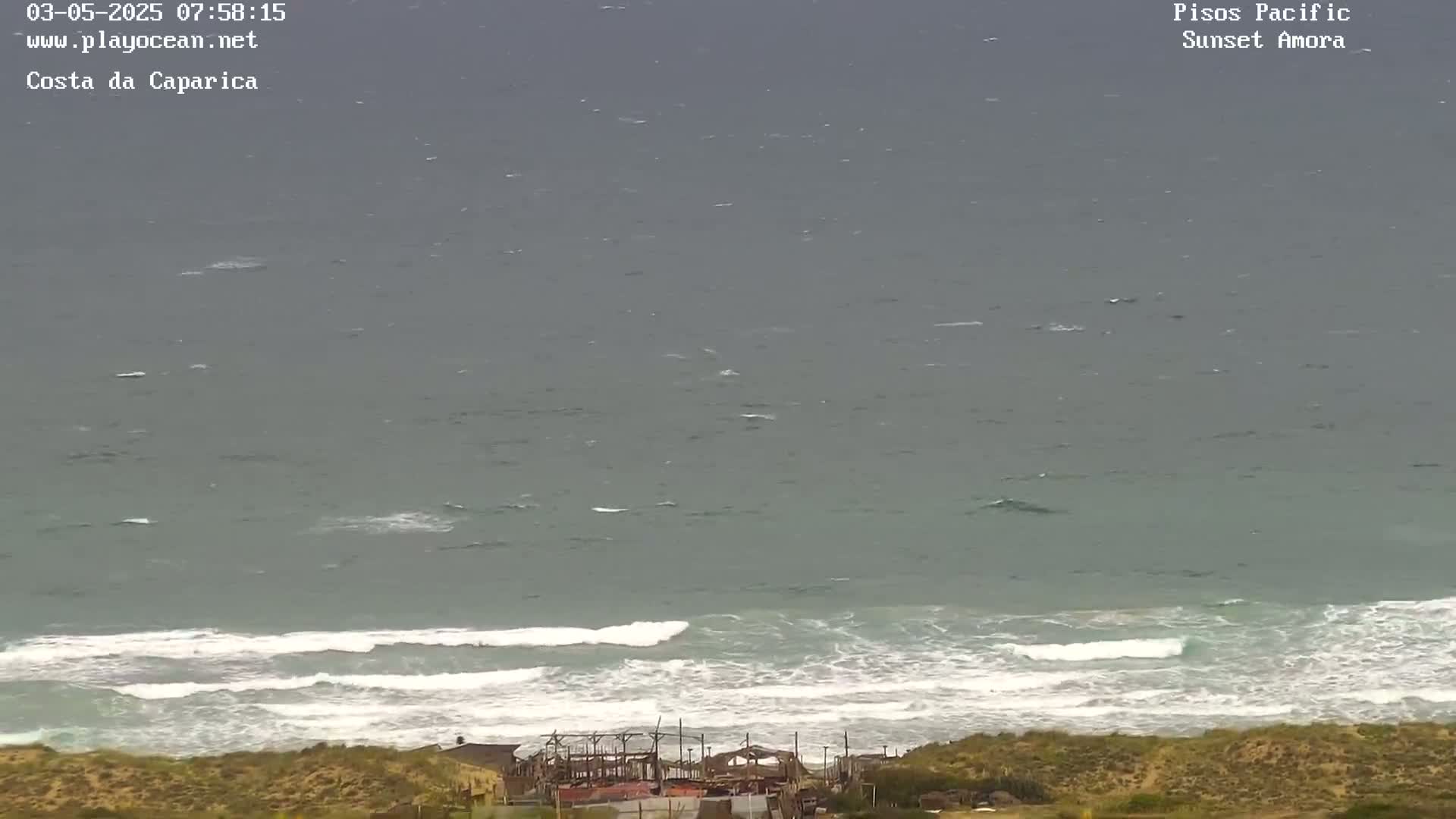 A high-angle view shows a grey ocean with moderate waves breaking onto a sandy shore, where some structures are visible.