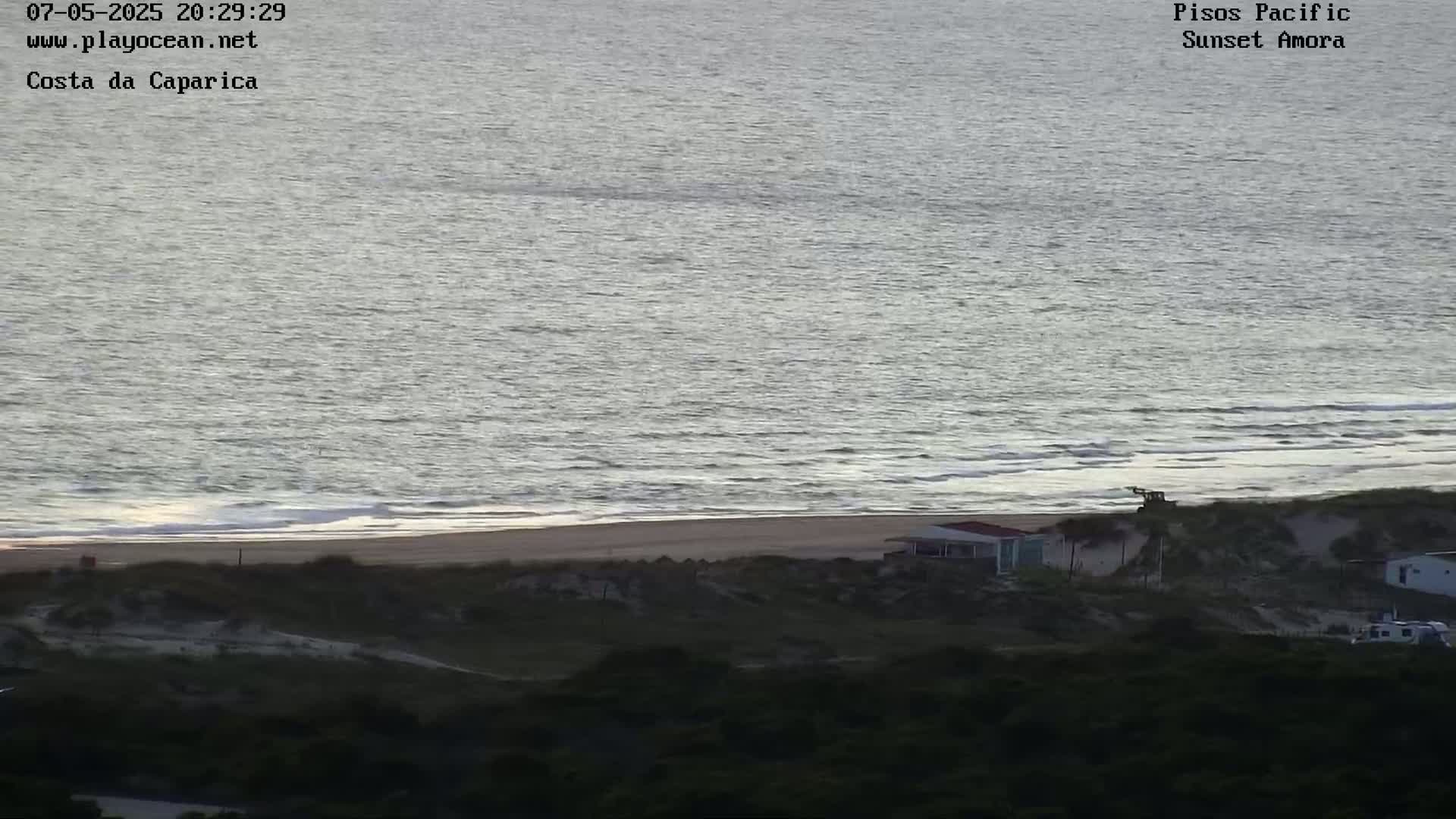 A beach with calm, gray water and a small building near the shoreline is seen from a hilltop, under a dim, overcast sky.