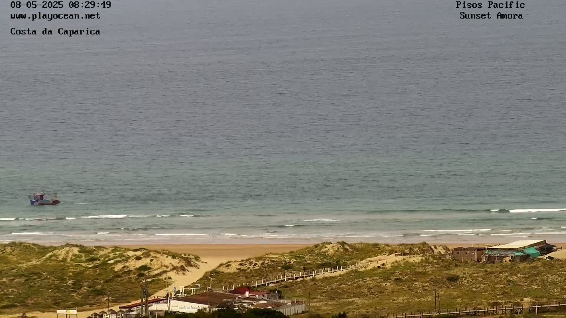 A small fishing boat is visible on a calm ocean with gentle waves breaking on a sandy beach backed by sand dunes and some low buildings.