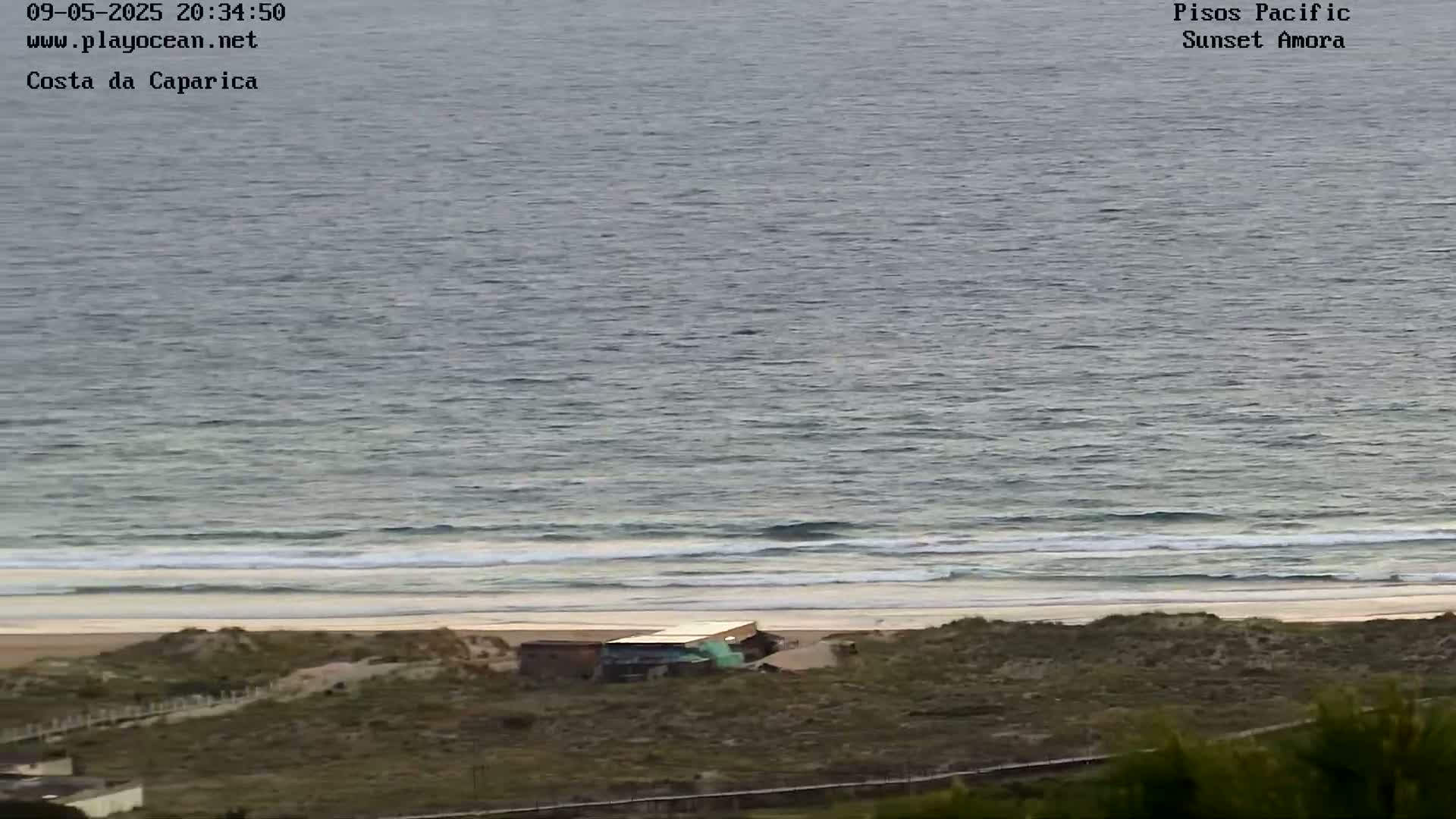 A calm ocean with small waves laps a sandy beach, where a small building sits behind a dune; the overall setting is somewhat overcast.