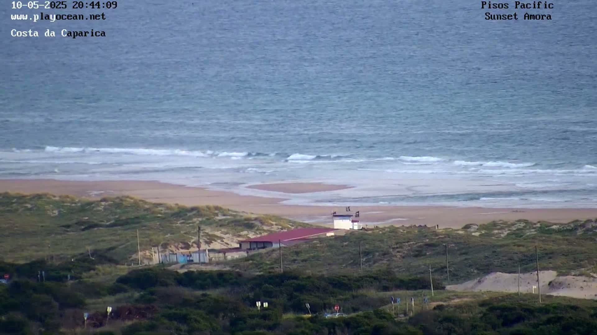 A sandy beach with gentle waves on a calm ocean is seen from an elevated vantage point, showing a small building and surrounding vegetation.