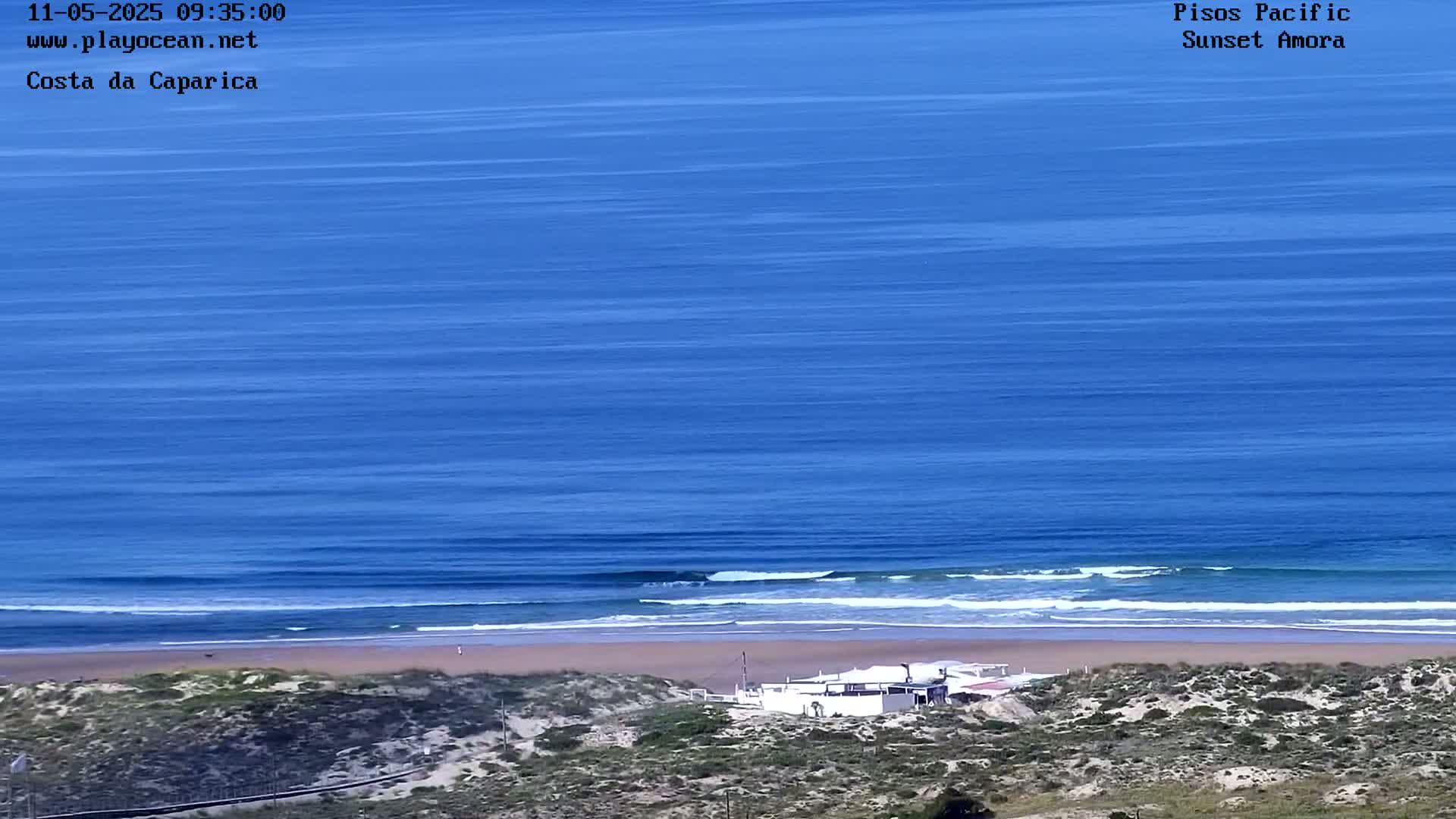 A sandy beach with a small building is seen from above, next to a blue ocean with small waves under a clear sky.