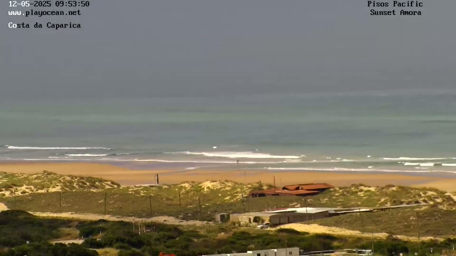 A wide, long shot shows a beach with gentle waves under an overcast sky, featuring buildings and sand dunes in the foreground.