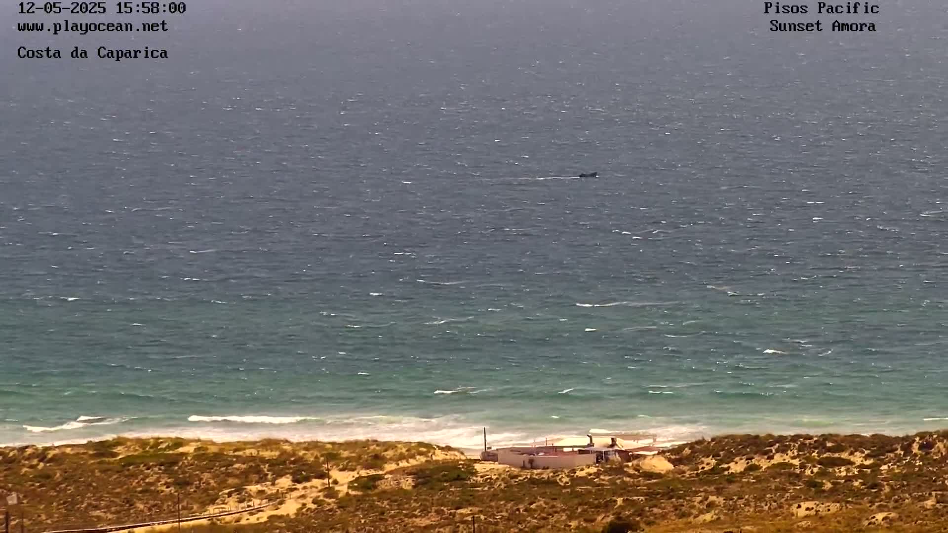 A small boat is visible on a choppy ocean, near a sandy beach with a small building, under a mostly cloudy sky.