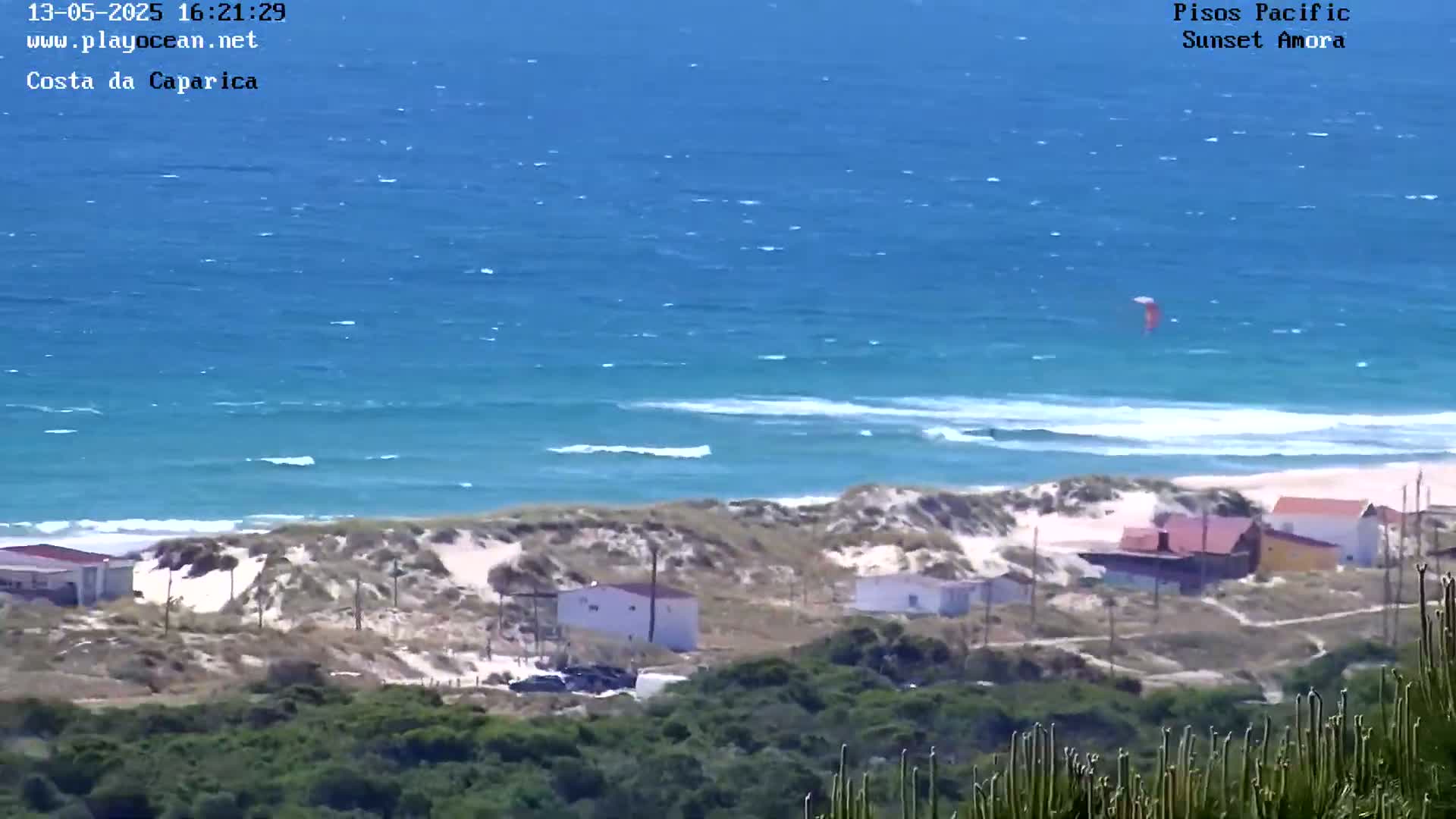 A kitesurfer is visible in the distance over a wavy ocean next to a sandy beach with a few buildings and vegetation.