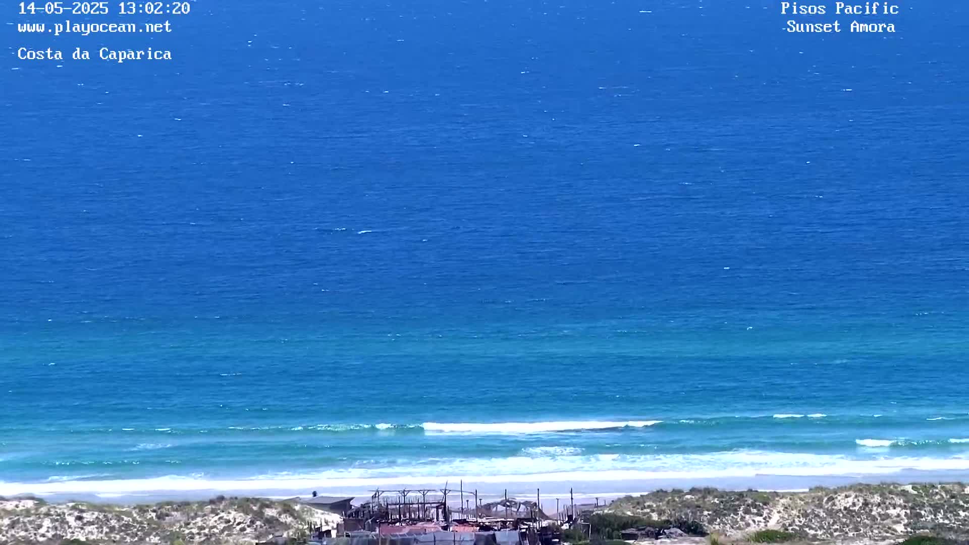 A high-angle view shows a vast, blue ocean with gentle waves rolling onto a sandy beach, where several structures are visible near the shore under a clear sky.