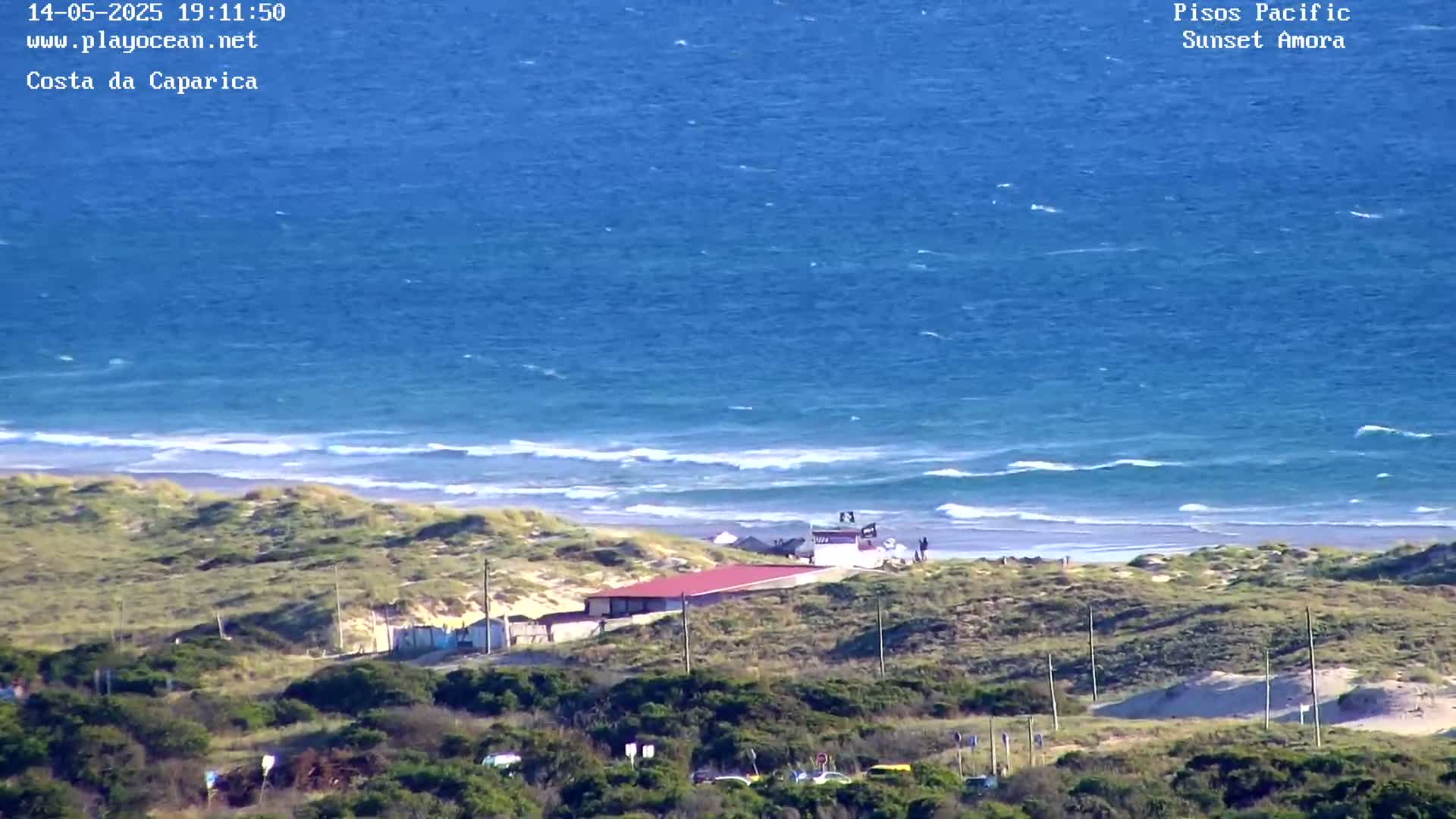 A high-angle view shows a sandy beach with a small building near a dune, overlooking a blue ocean with small waves under a mostly sunny sky.