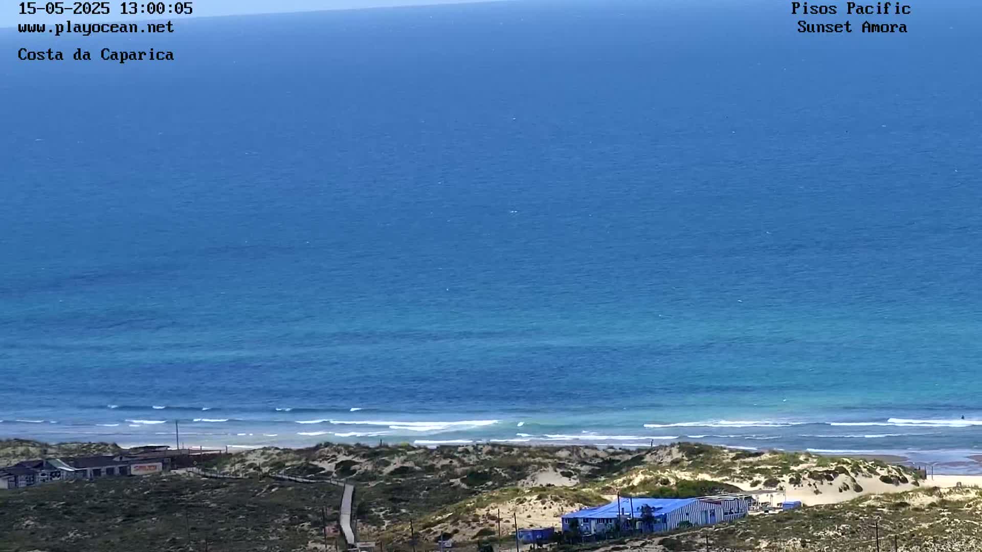 A high-angle view shows a mostly clear, blue ocean with small waves lapping a sandy shore, backed by low-lying vegetation and a few small buildings.
