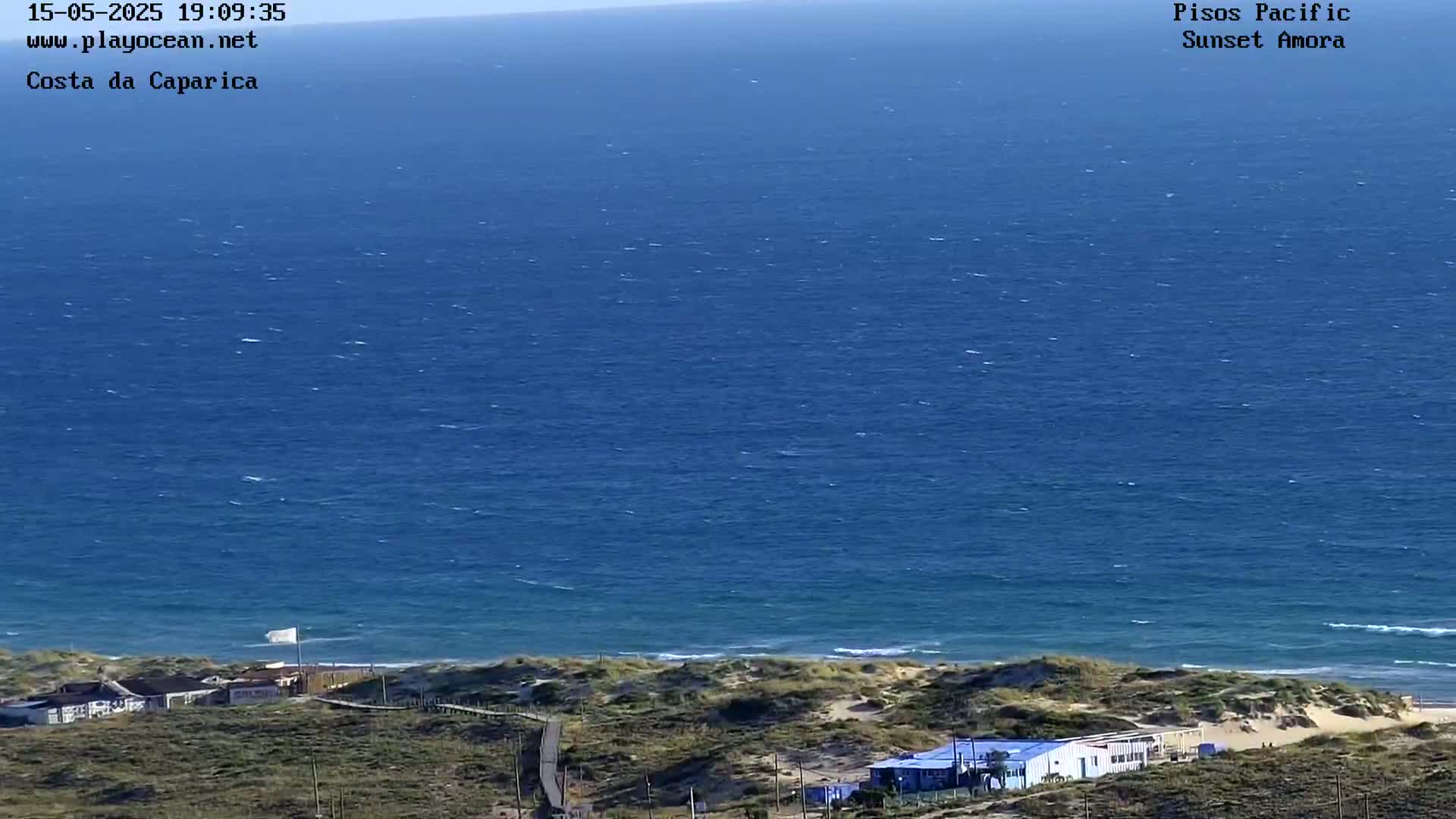 A coastal view shows a wide expanse of blue ocean with small waves, a sandy beach, and low-lying buildings with vegetation in the foreground under a clear sky.