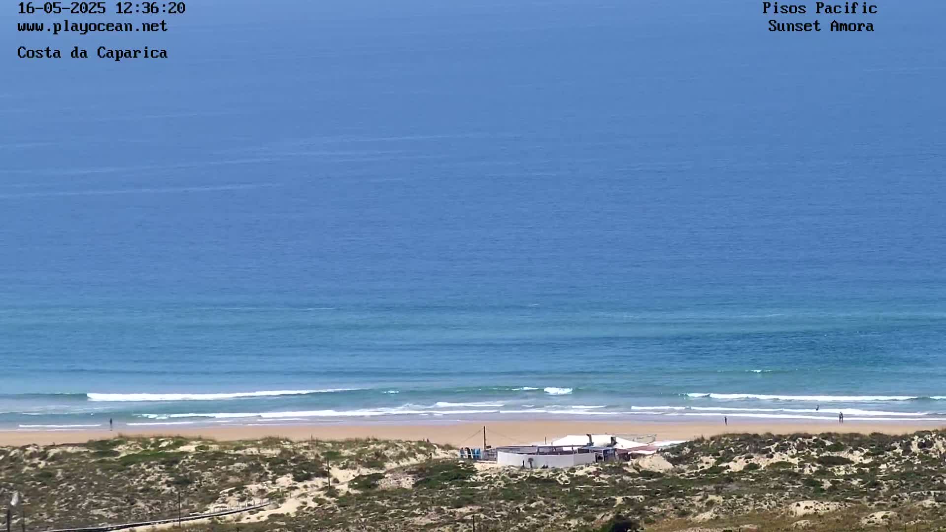 A sandy beach with a small building and a few people is visible, overlooking a calm ocean under a clear blue sky.