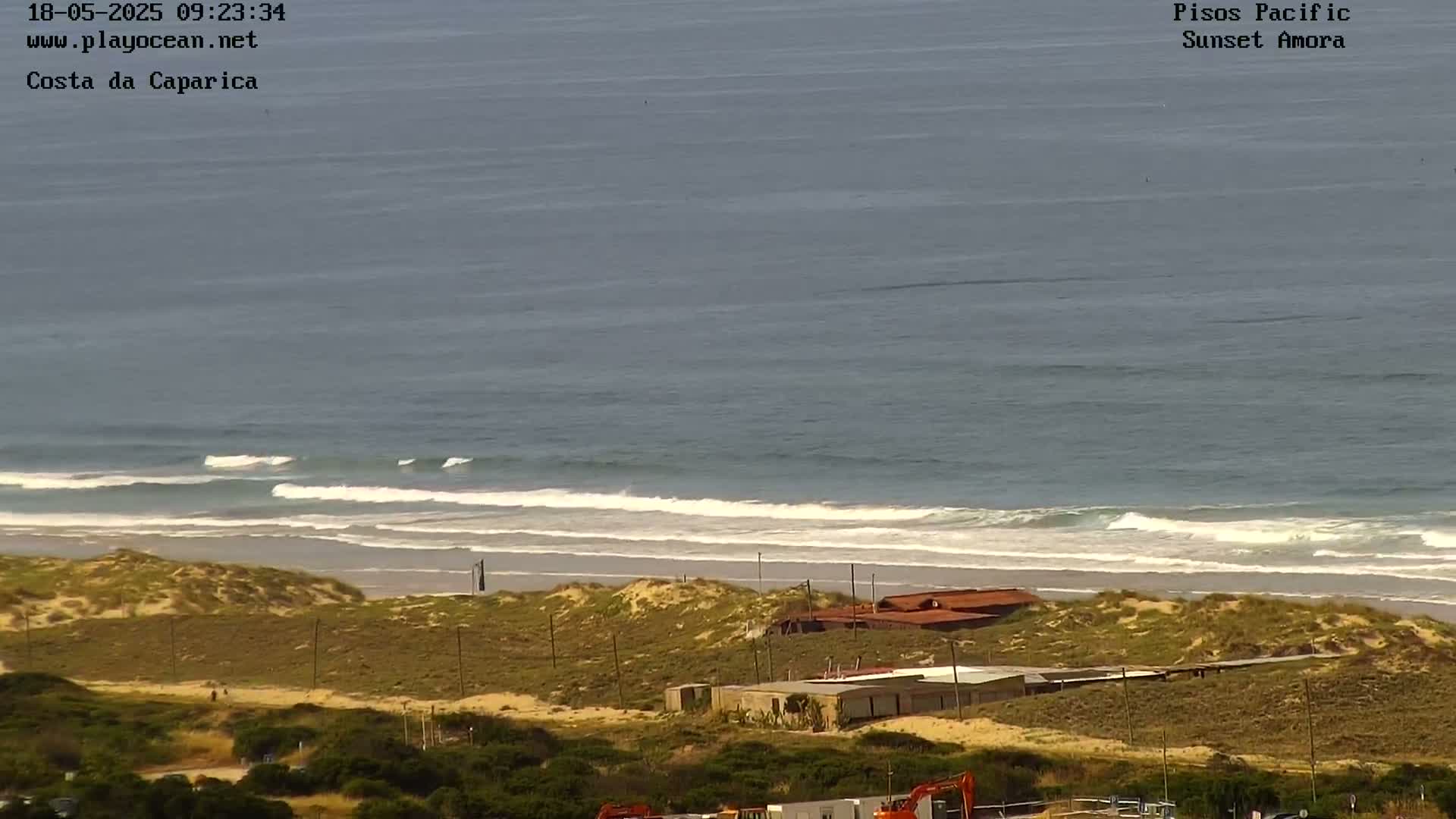 A mostly sunny day at a beach shows small waves rolling onto the shore, with sand dunes and buildings visible beyond the beach.