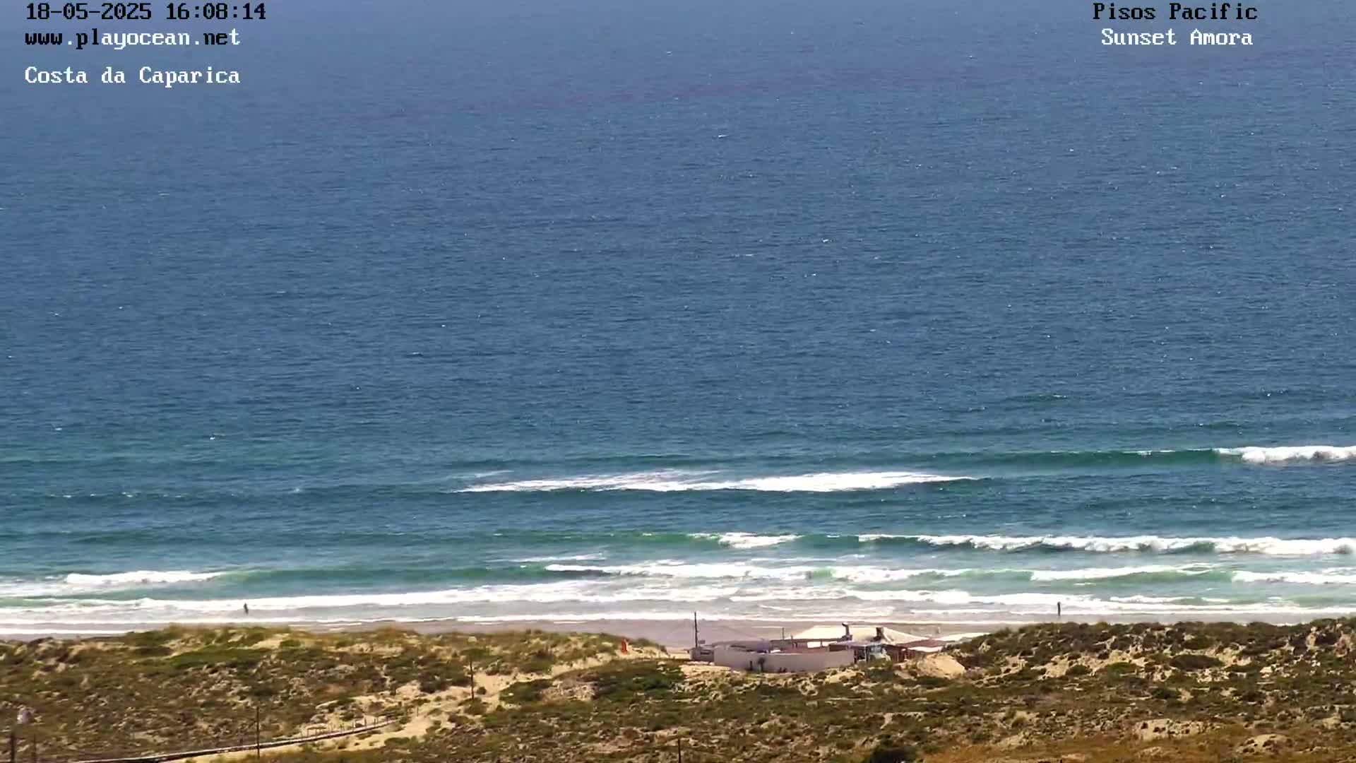 A sandy beach with a small building is shown, with ocean waves rolling in under a clear sky.