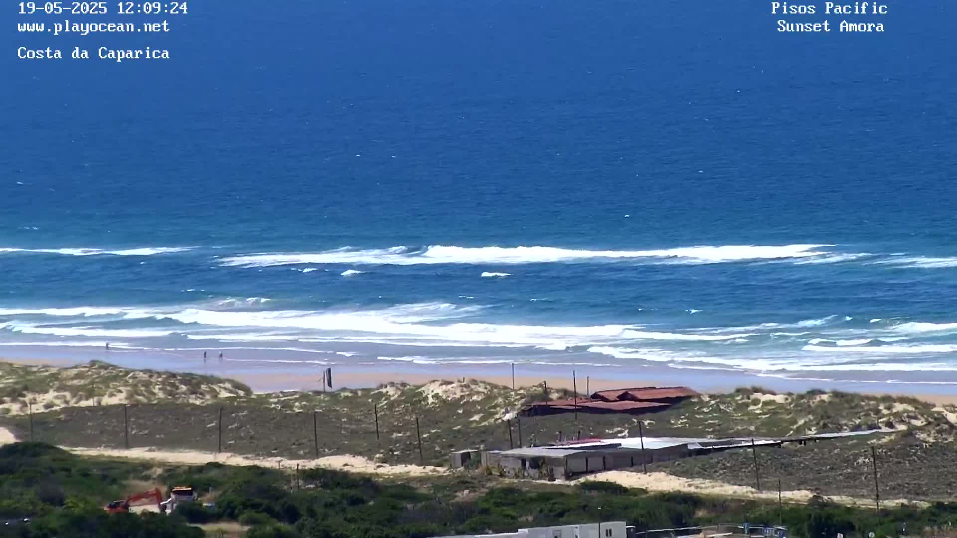 A sandy beach with small waves rolling onto the shore is backed by sand dunes, sparse vegetation, and a few low-lying buildings, all under a clear, sunny sky.