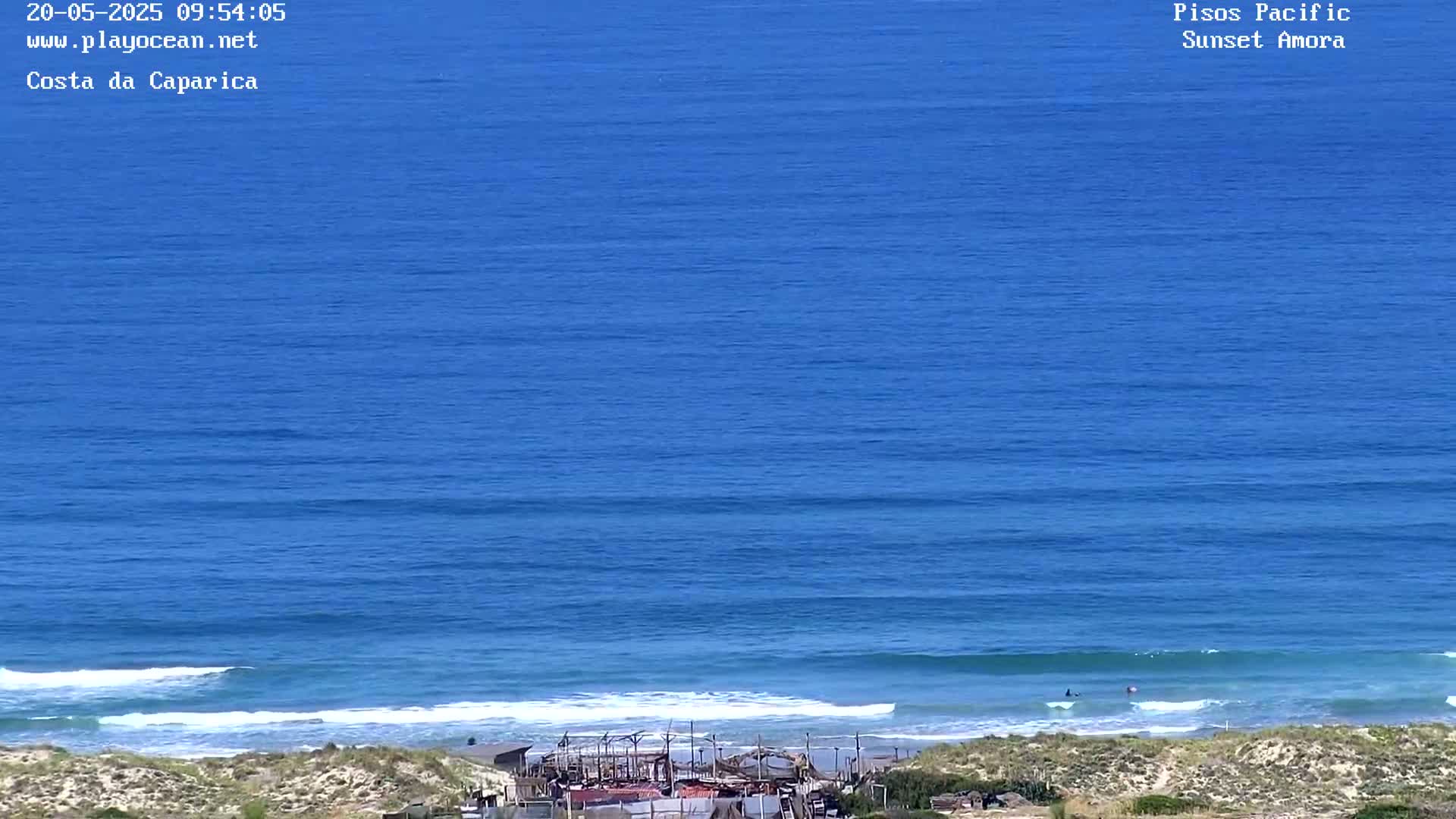A mostly sunny day shows a wide expanse of deep blue ocean with small waves rolling onto a sandy beach, where some structures are visible near the shoreline.