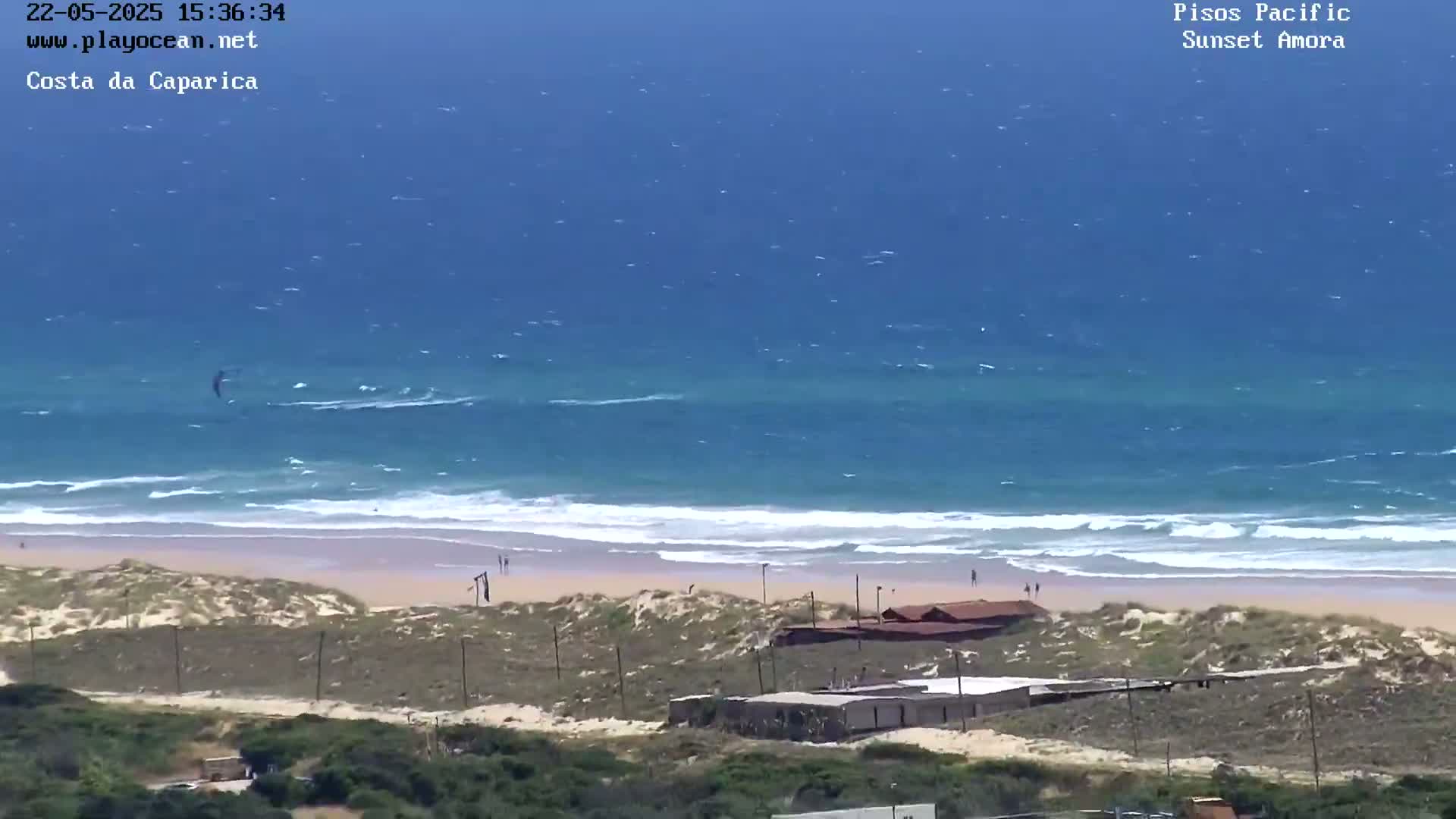 A kitesurfer is visible in the ocean near a sandy beach with some buildings and vegetation in the foreground under sunny conditions.