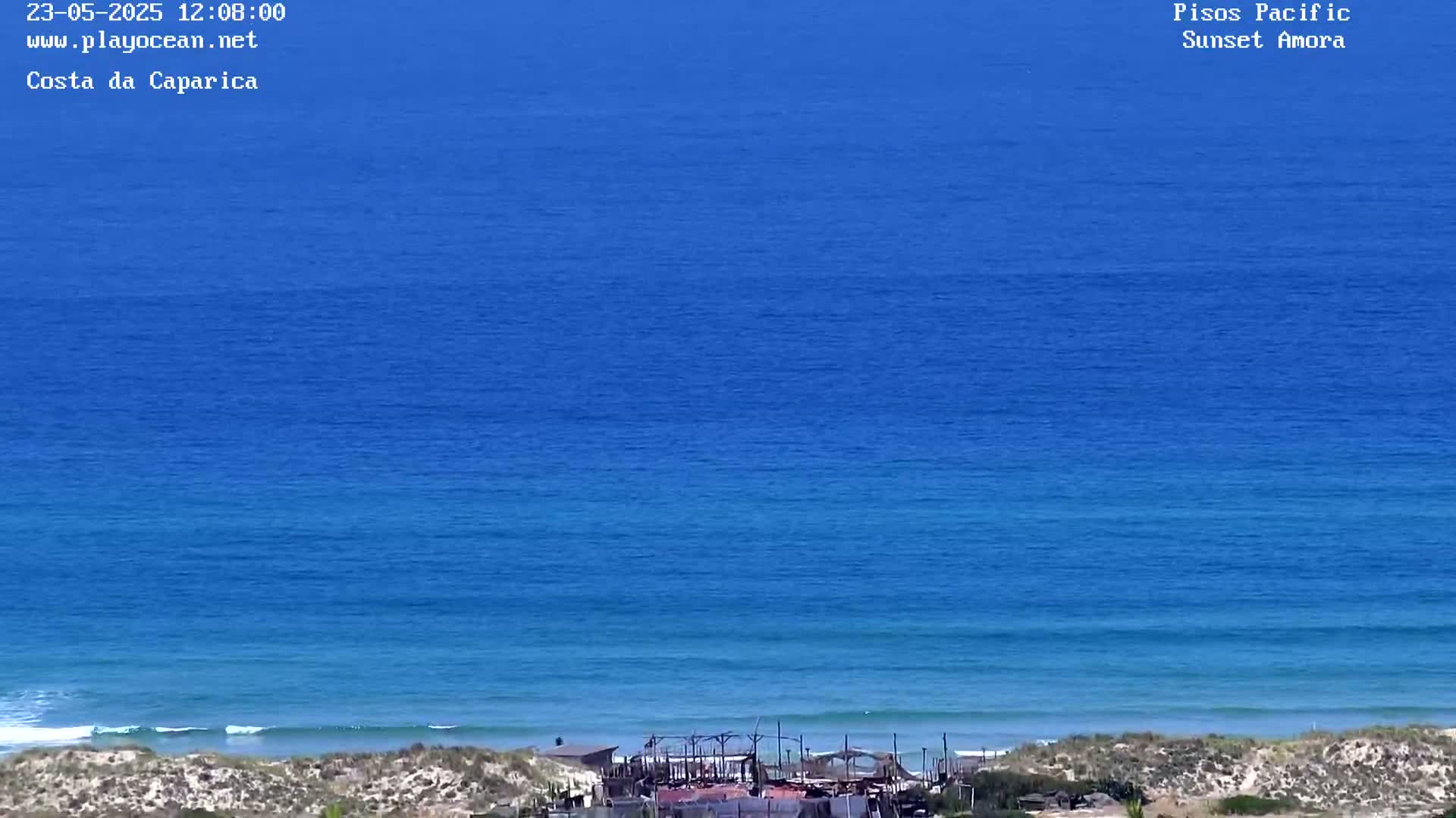 A mostly sunny day shows a wide expanse of deep blue ocean meeting a sandy shore with some structures visible near the waterline.