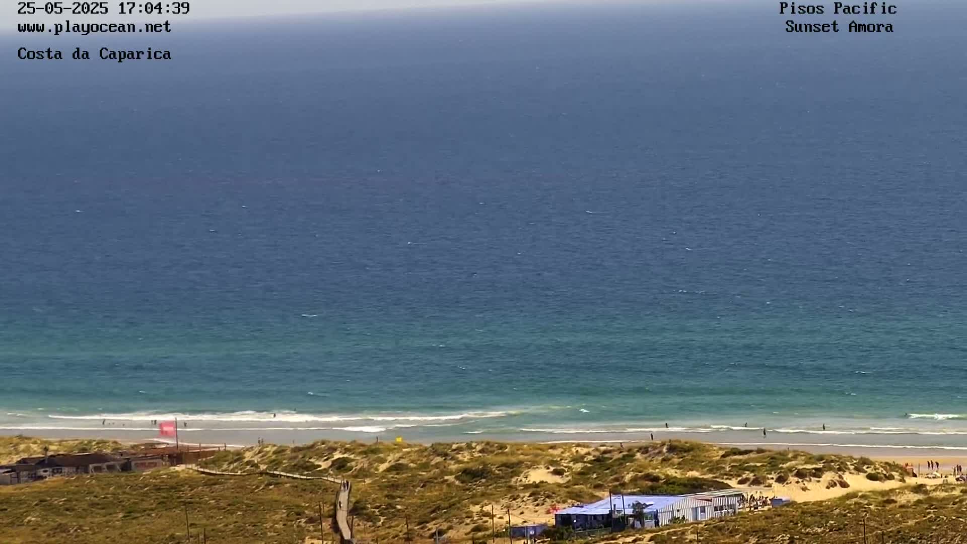 An ocean beach with calm waves and a sandy shore is seen from an elevated viewpoint on a sunny day.