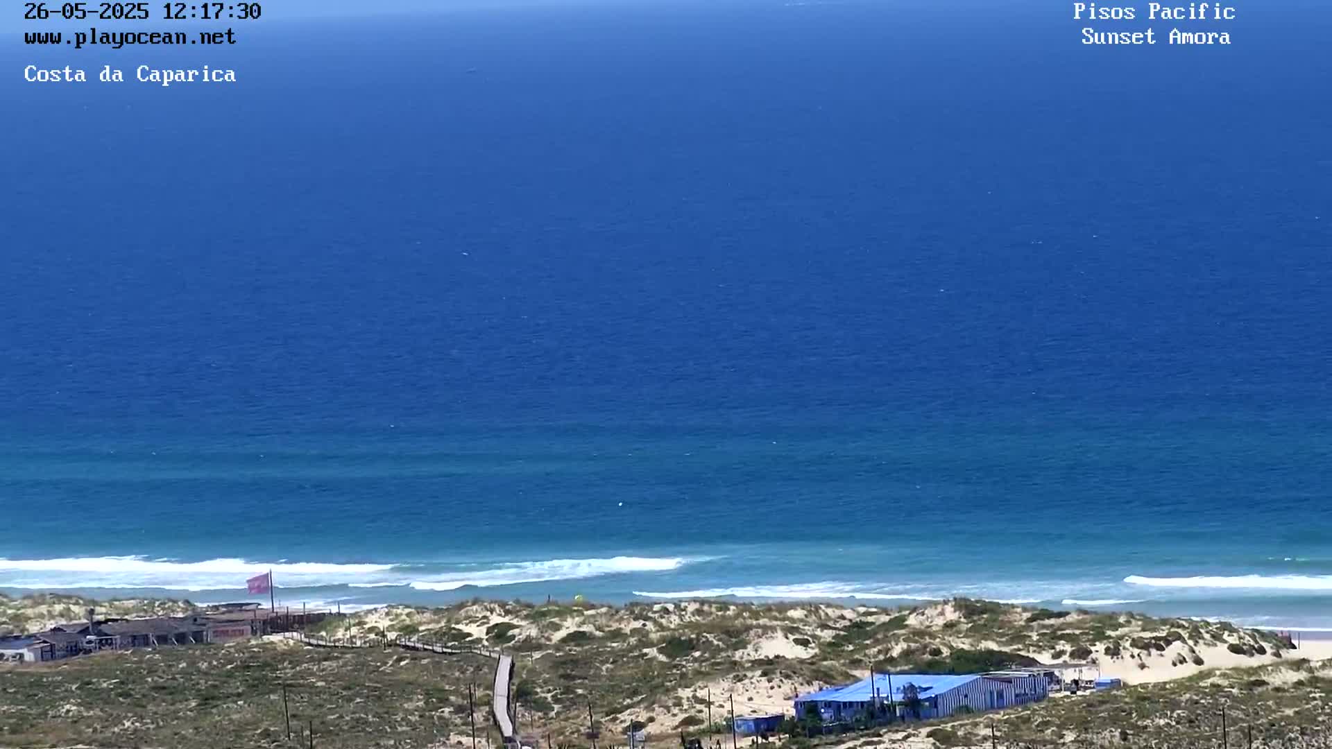 A high-angle, long shot shows a mostly sunny day at a sandy beach with light waves lapping the shore and a calm, blue ocean extending to the horizon.