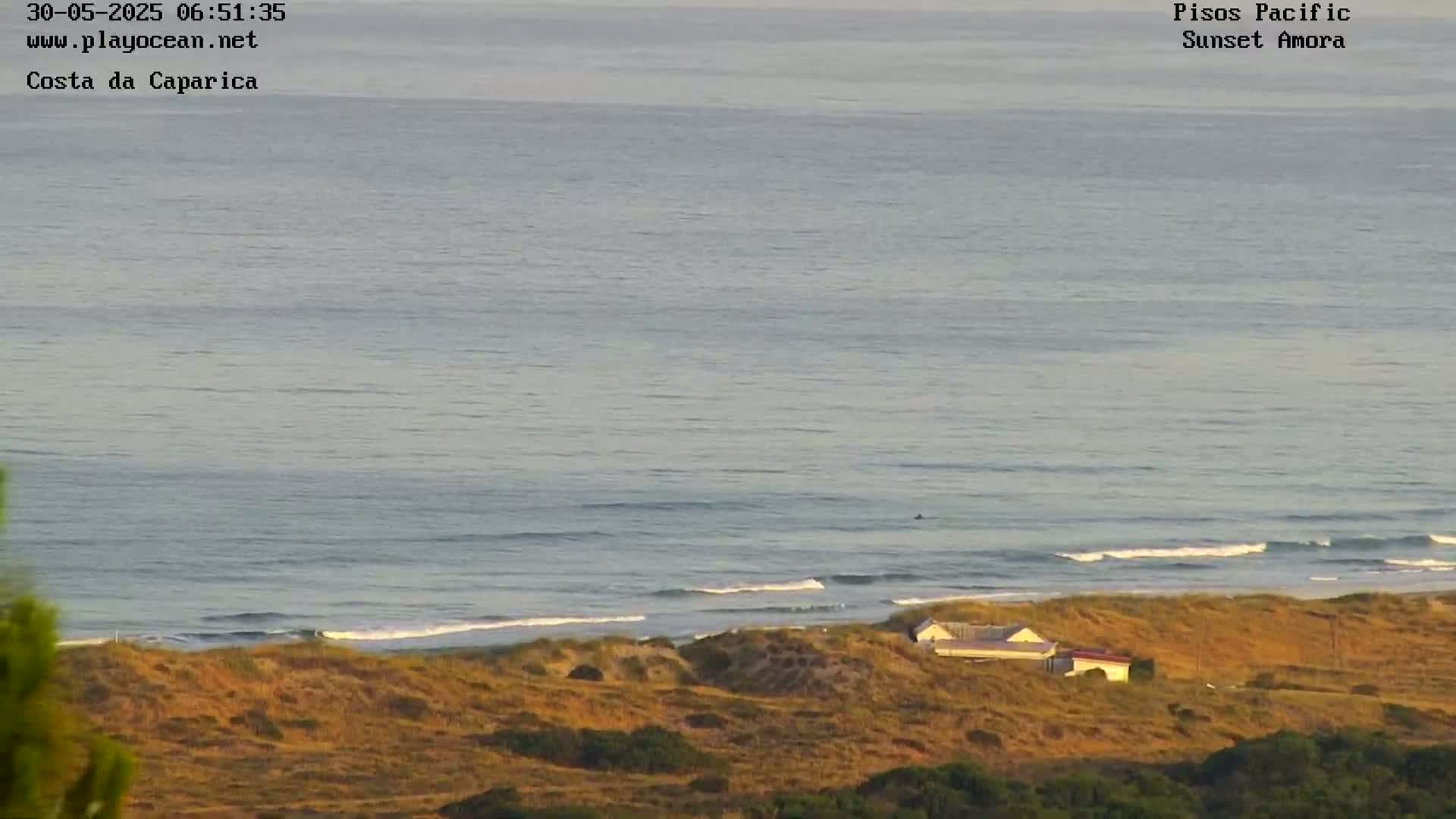 A calm ocean with small waves laps a sandy shore where a single building sits atop a dry, golden-brown hillside under a mostly clear sky.