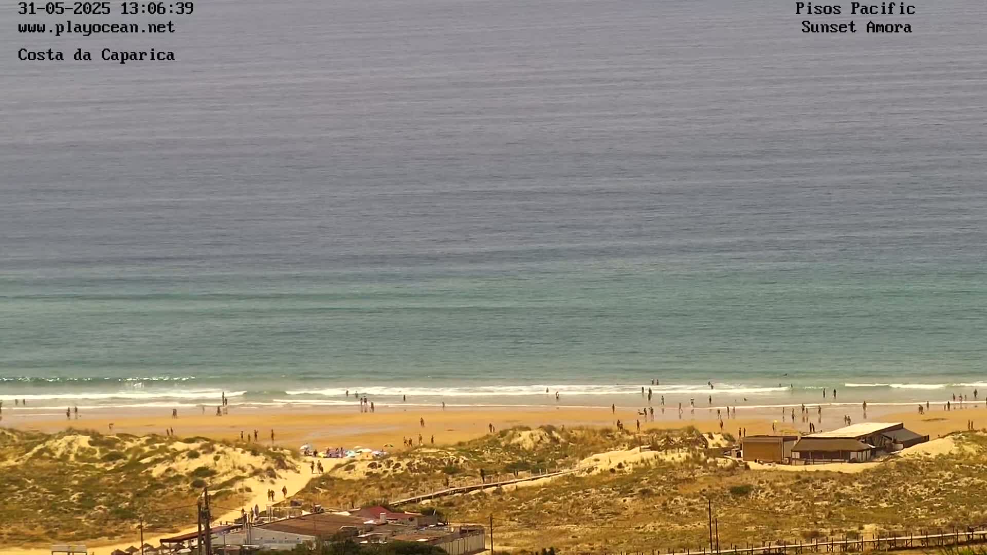 An aerial view shows a sandy beach with people scattered along the shore and in the water, under a mostly clear sky, with calm ocean waves breaking gently near the beach.