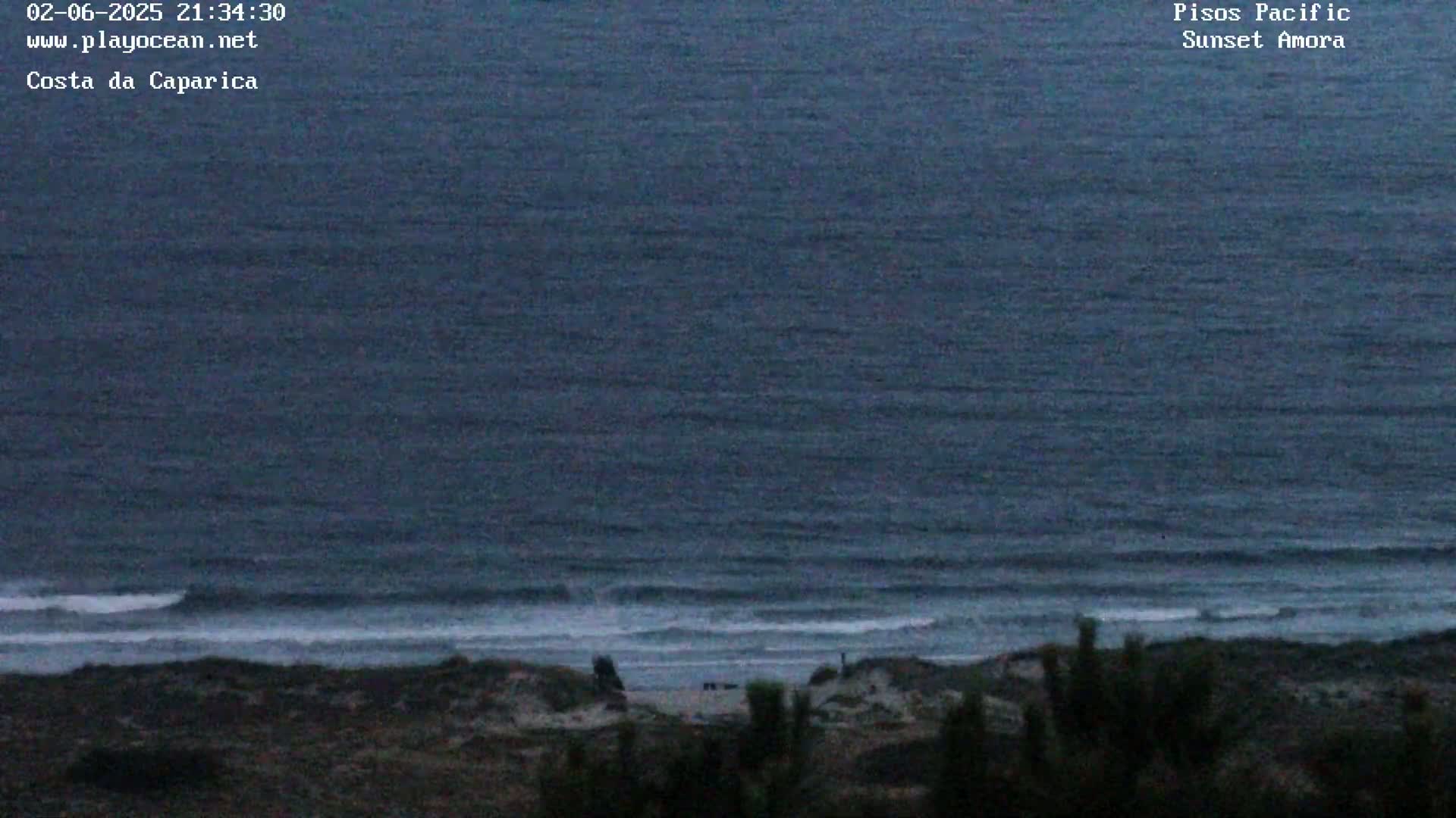 A dark blue ocean with small waves rolls onto a dark, sandy shore under a twilight sky.