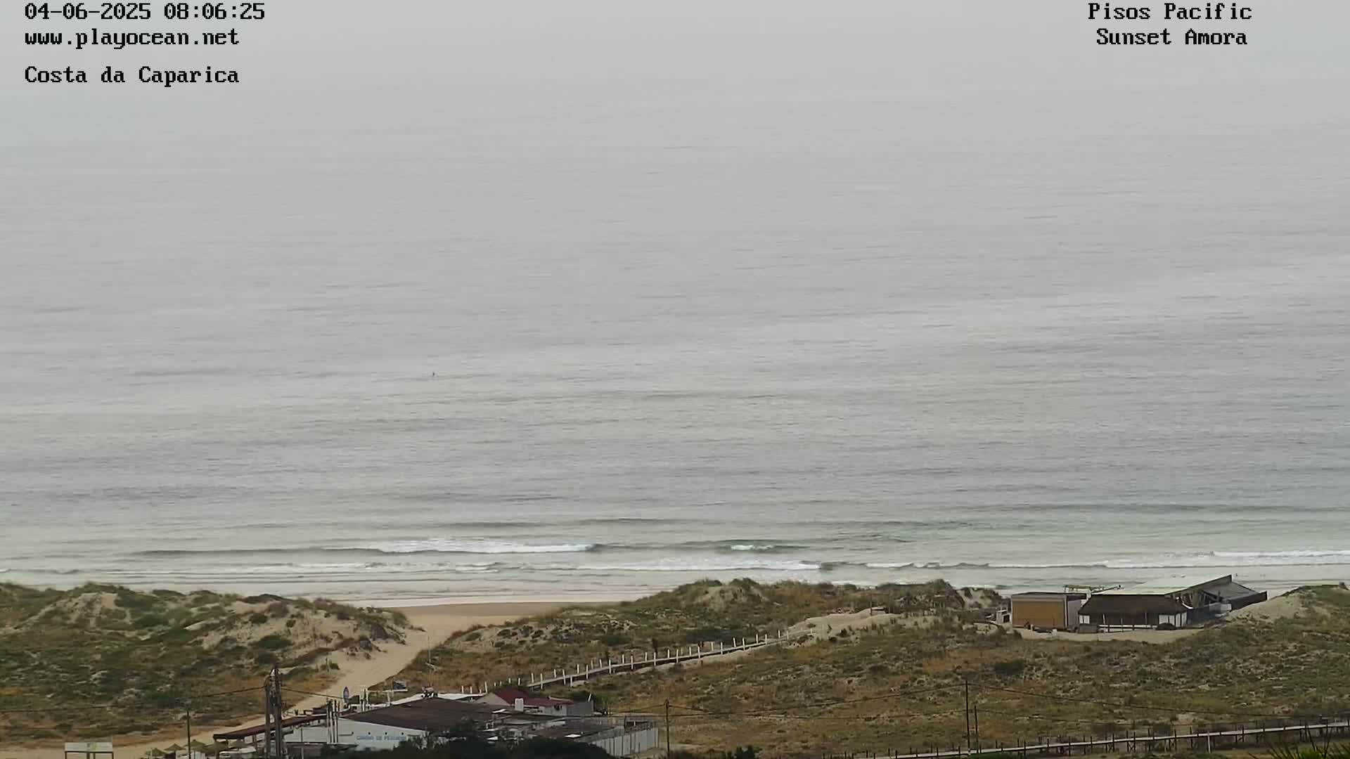 A hazy view shows an ocean beach with small waves lapping a sandy shore, backed by sand dunes and a few buildings.