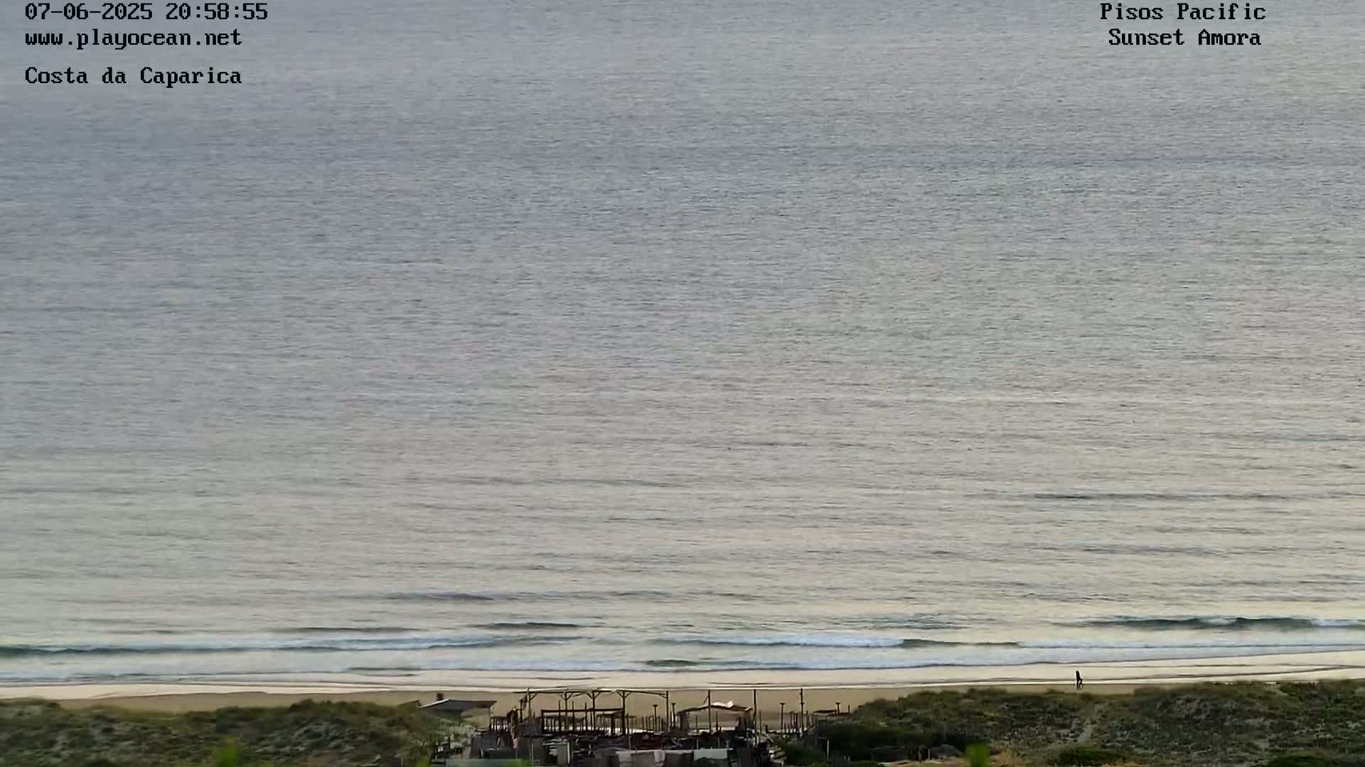 A long shot of a calm ocean meeting a sandy beach with small structures visible near the shore under a muted sky.