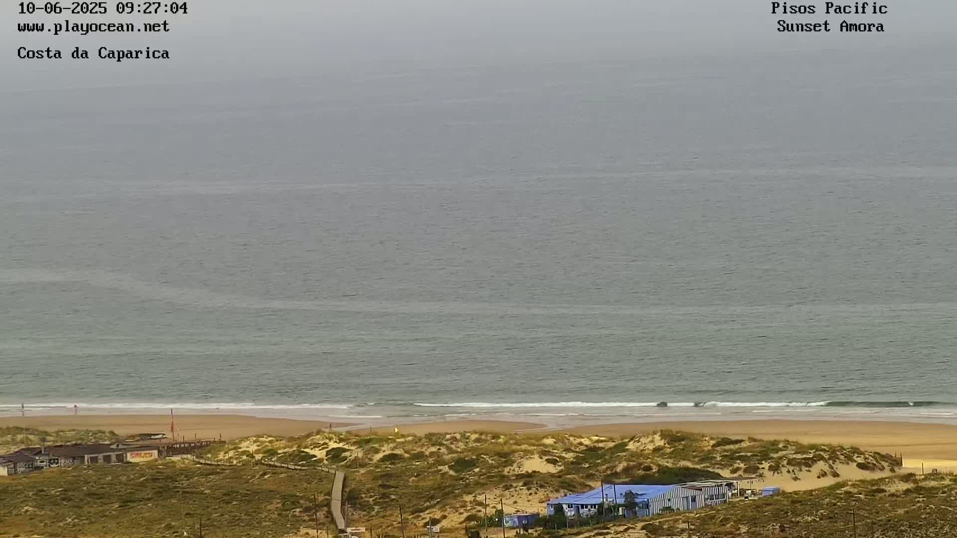 A hazy, overcast day reveals a beach with sand dunes and a few buildings, overlooking a calm ocean.
