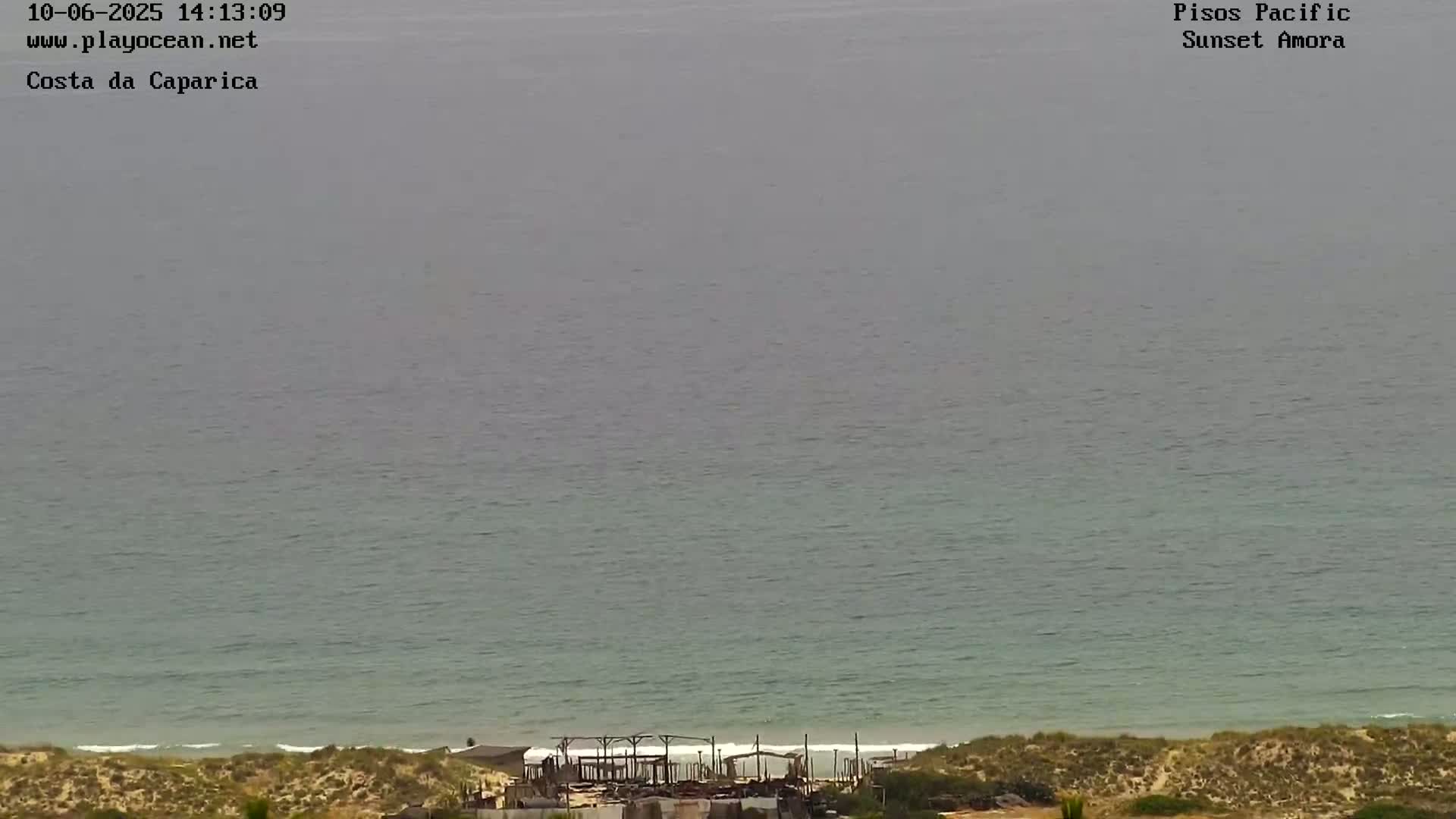 A mostly overcast view shows a calm ocean meeting a sandy beach with a structure near the shoreline.