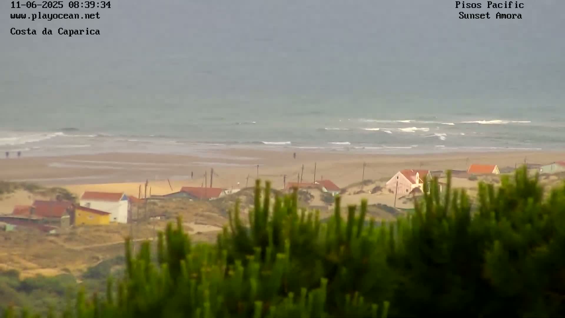 A view from a dune shows a beach with small waves under an overcast sky, behind which are small houses nestled in sand dunes.