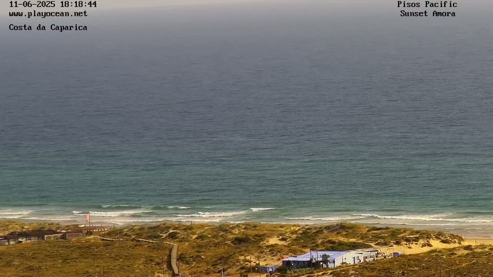 A mostly clear, hazy day shows a wide expanse of ocean meeting a sandy beach with small buildings and a boardwalk.