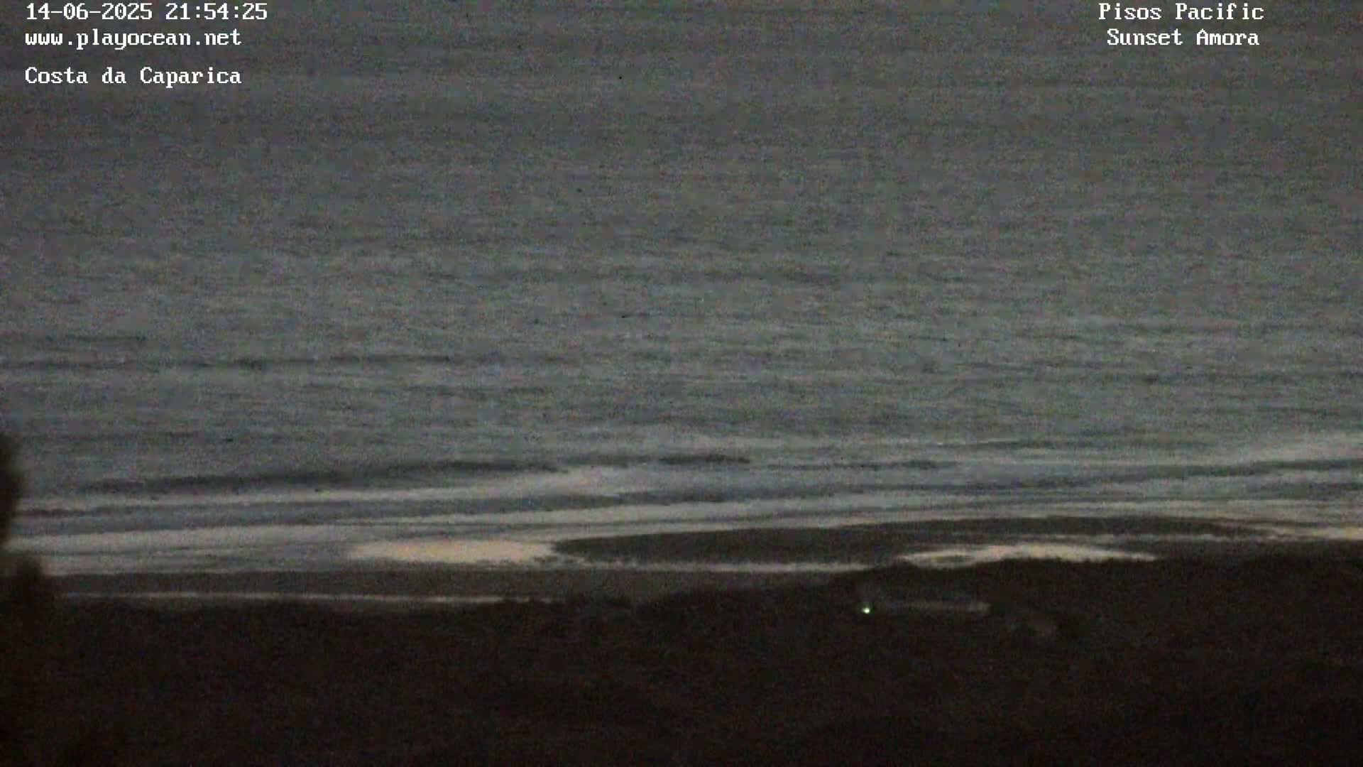 A mostly clear, hazy day shows a wide expanse of ocean meeting a sandy beach with small buildings and a boardwalk.