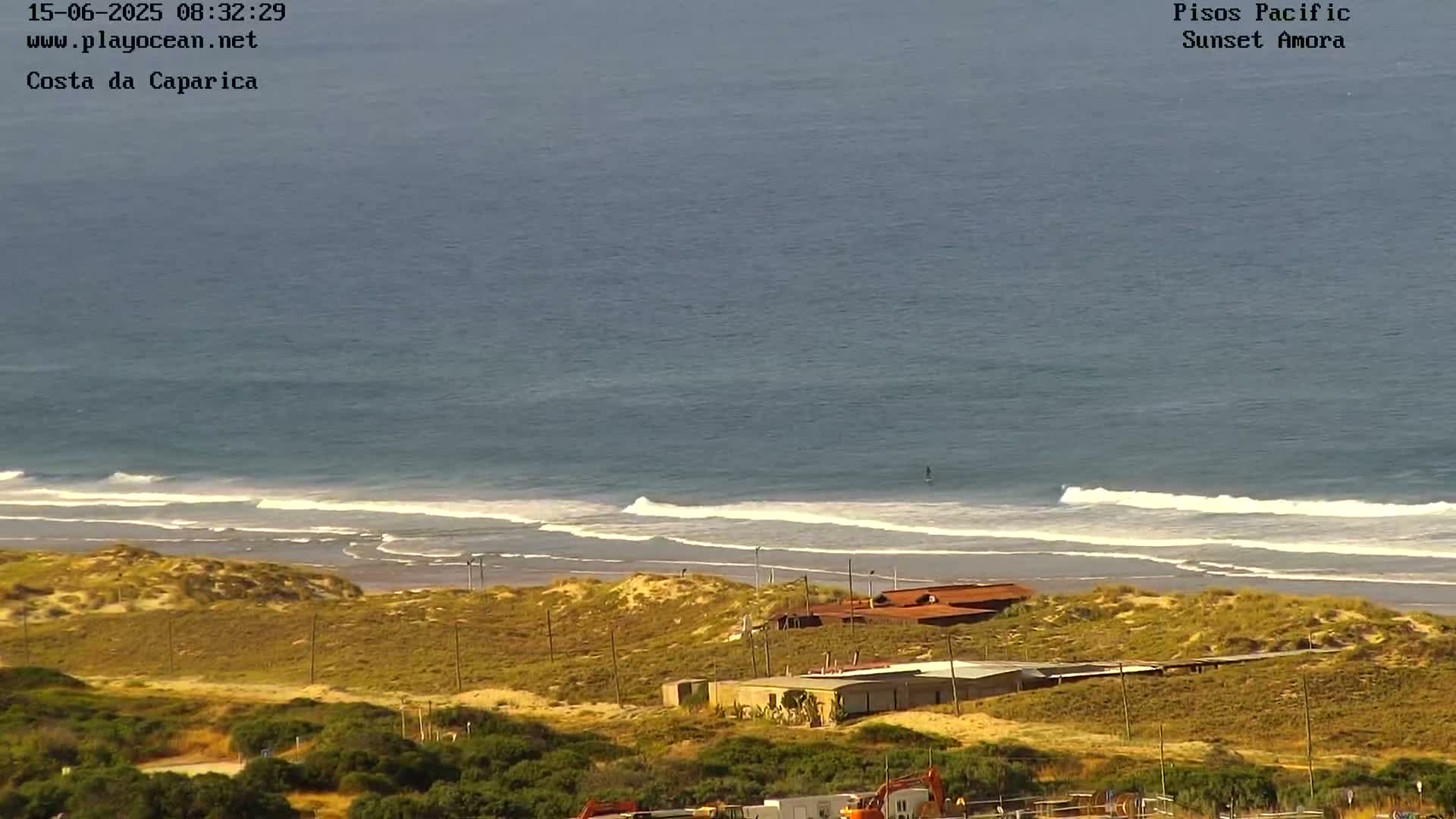 A high-angle view shows a sandy beach with small waves lapping the shore, several buildings, and dry, shrub-covered dunes under a clear sky.