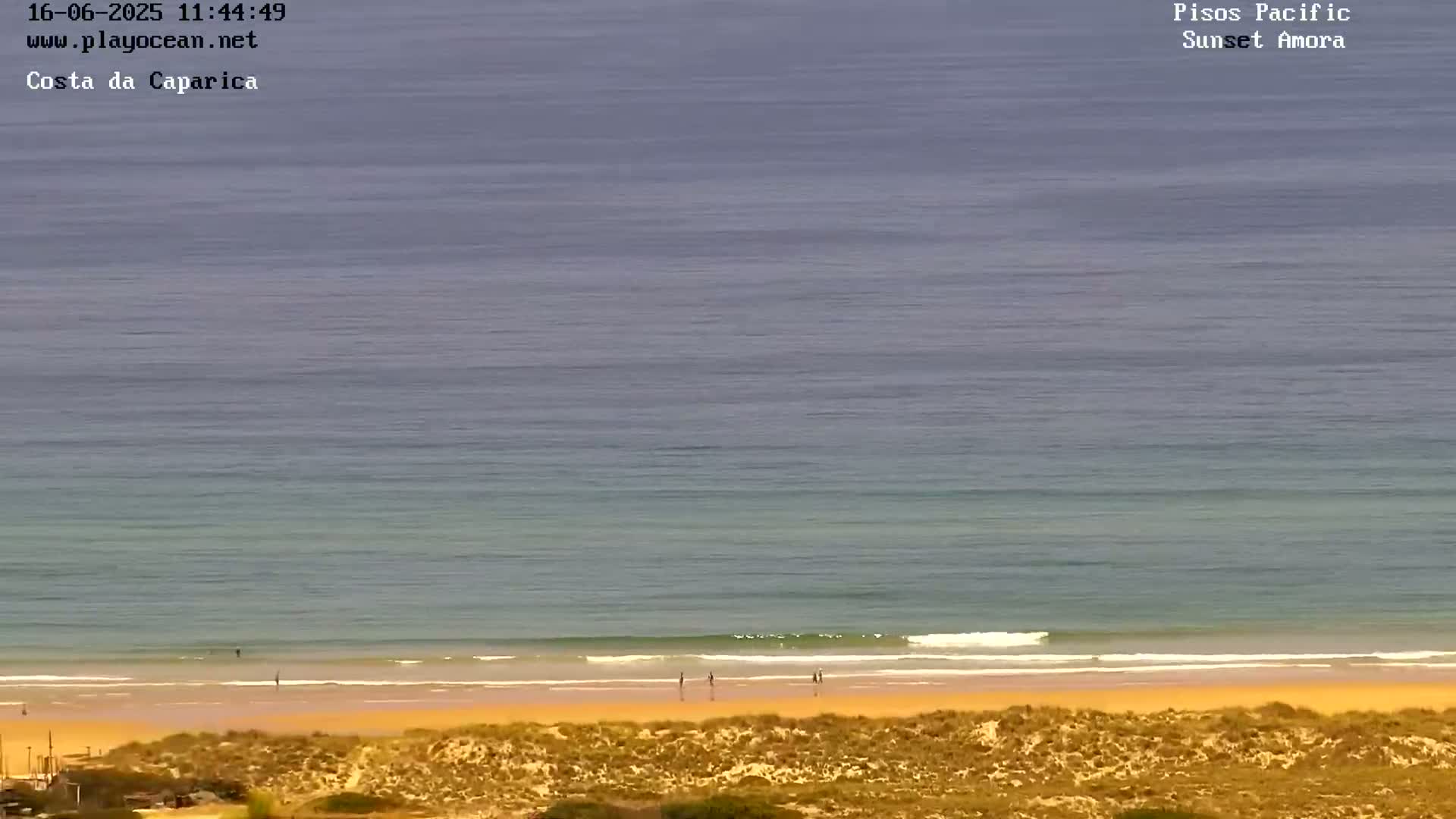 A mostly clear, sunny day shows a long expanse of ocean meeting a sandy beach with a few people visible near the water's edge.