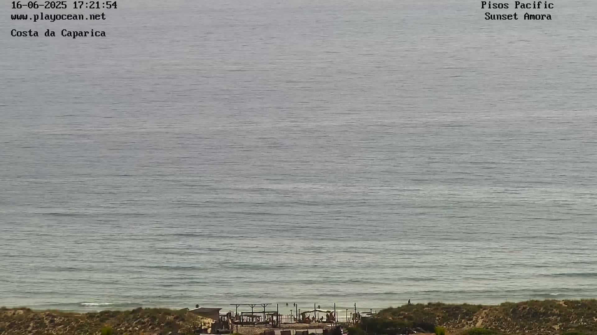A mostly calm, gray-green ocean stretches to the horizon, meeting a sandy shore with a few small structures visible in the distance.