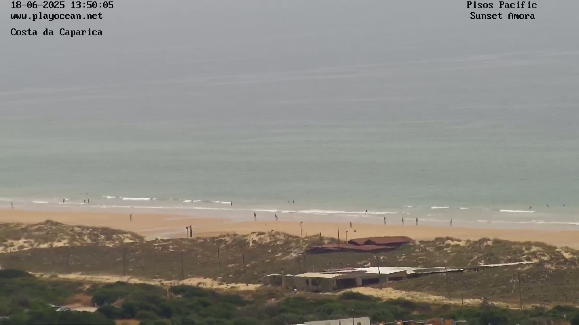 A hazy view shows a beach with a few people on it, backed by sand dunes and low buildings, all overlooking a calm, gray-green ocean.