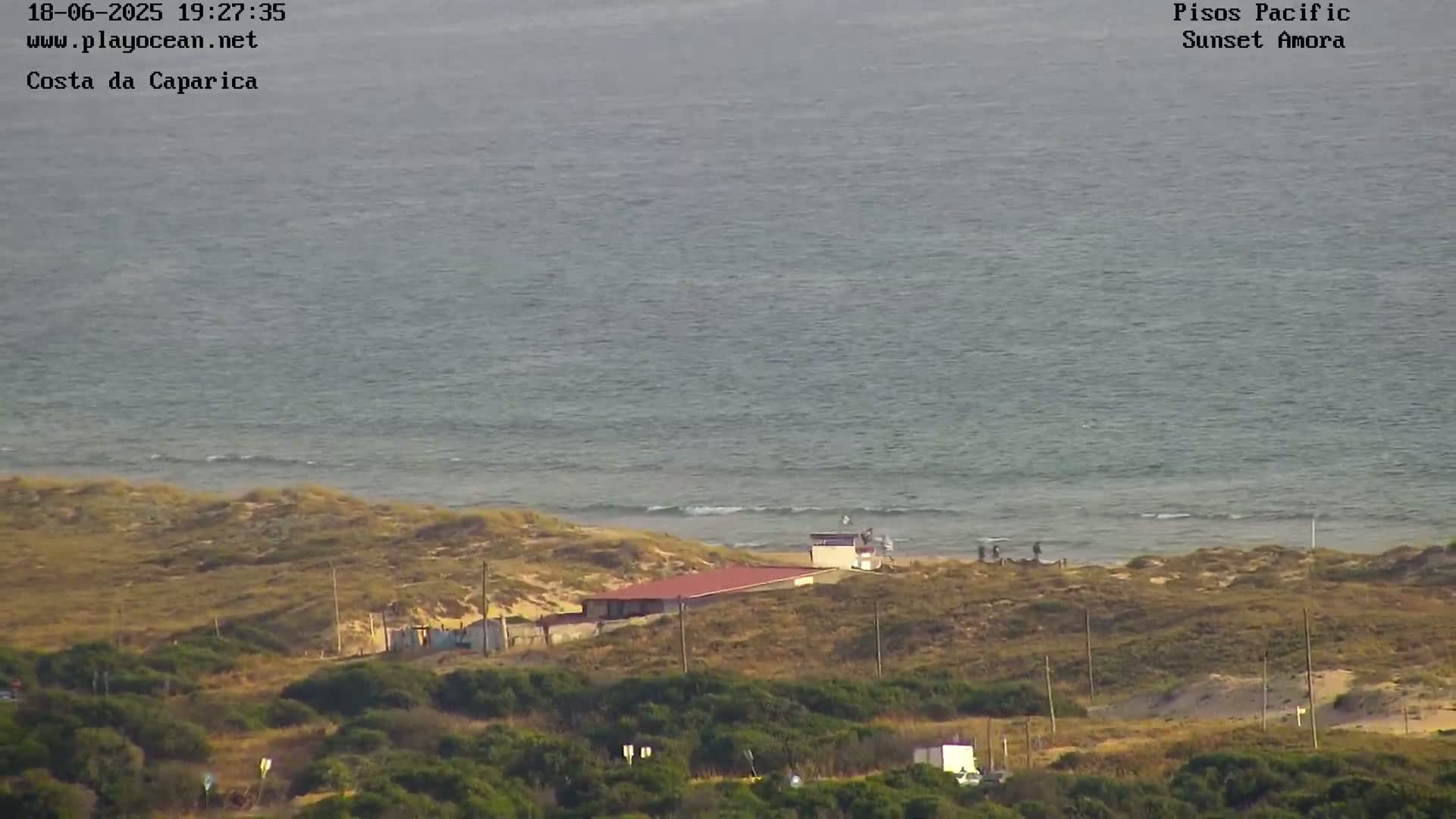 A beach with a calm ocean under a mostly clear sky features a low-lying building near the shoreline and vegetation covering the dunes.