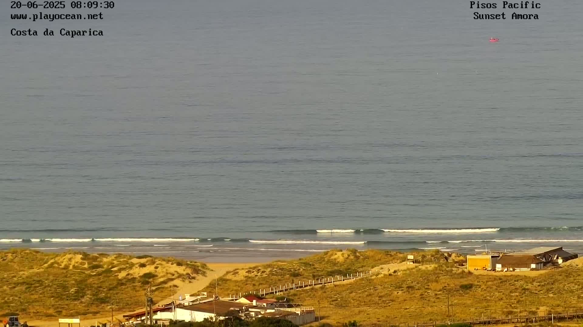 A calm ocean with small waves laps a sandy beach near some buildings under a mostly clear sky.