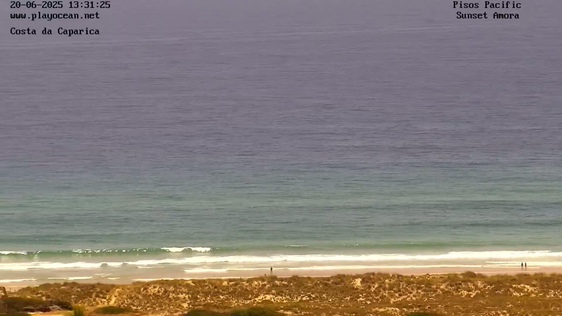A mostly clear, calm ocean meets a sandy beach backed by dry, tan dunes.