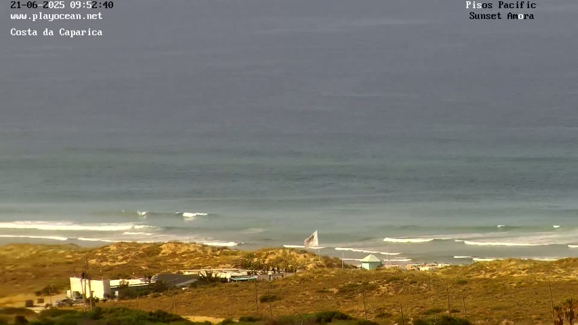 An overcast day shows a beach with small waves lapping the shore, a few buildings, and dry, sandy dunes.