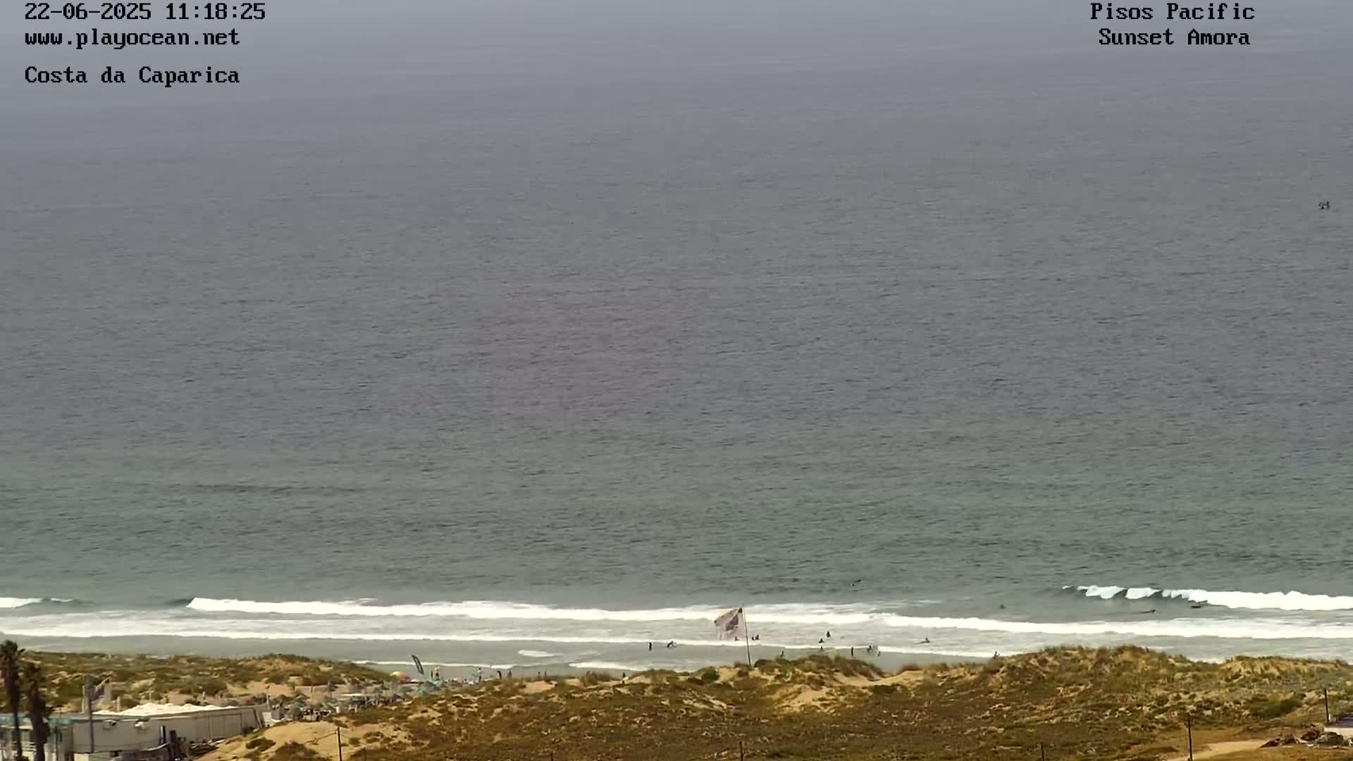 A mostly overcast day shows a view of a beach with several people near the water's edge, small waves breaking on the shore, and sand dunes in the foreground.