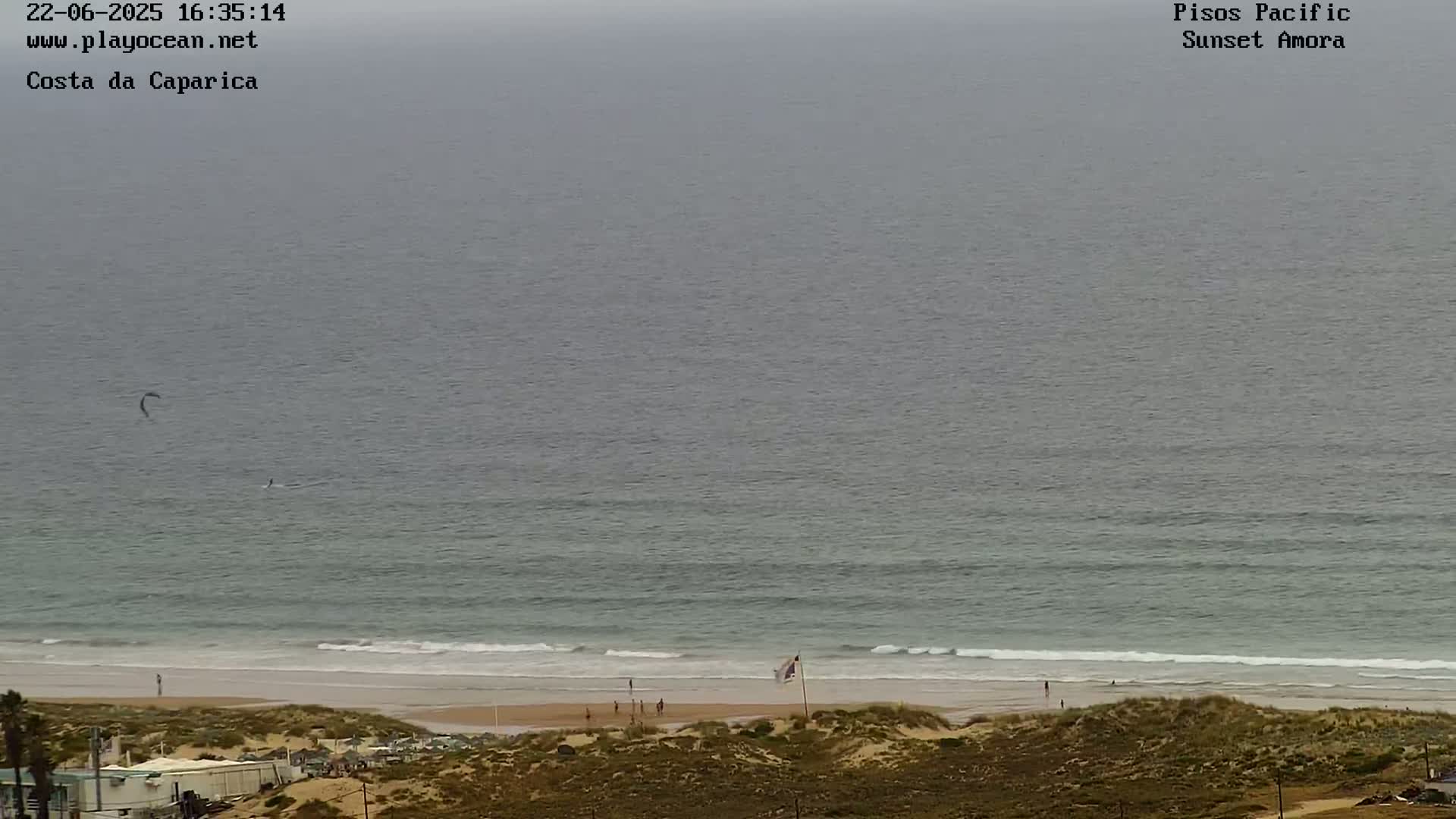 An overcast day shows a wide expanse of ocean meeting a sandy beach with a few people visible; a kitesurfer is seen in the water.