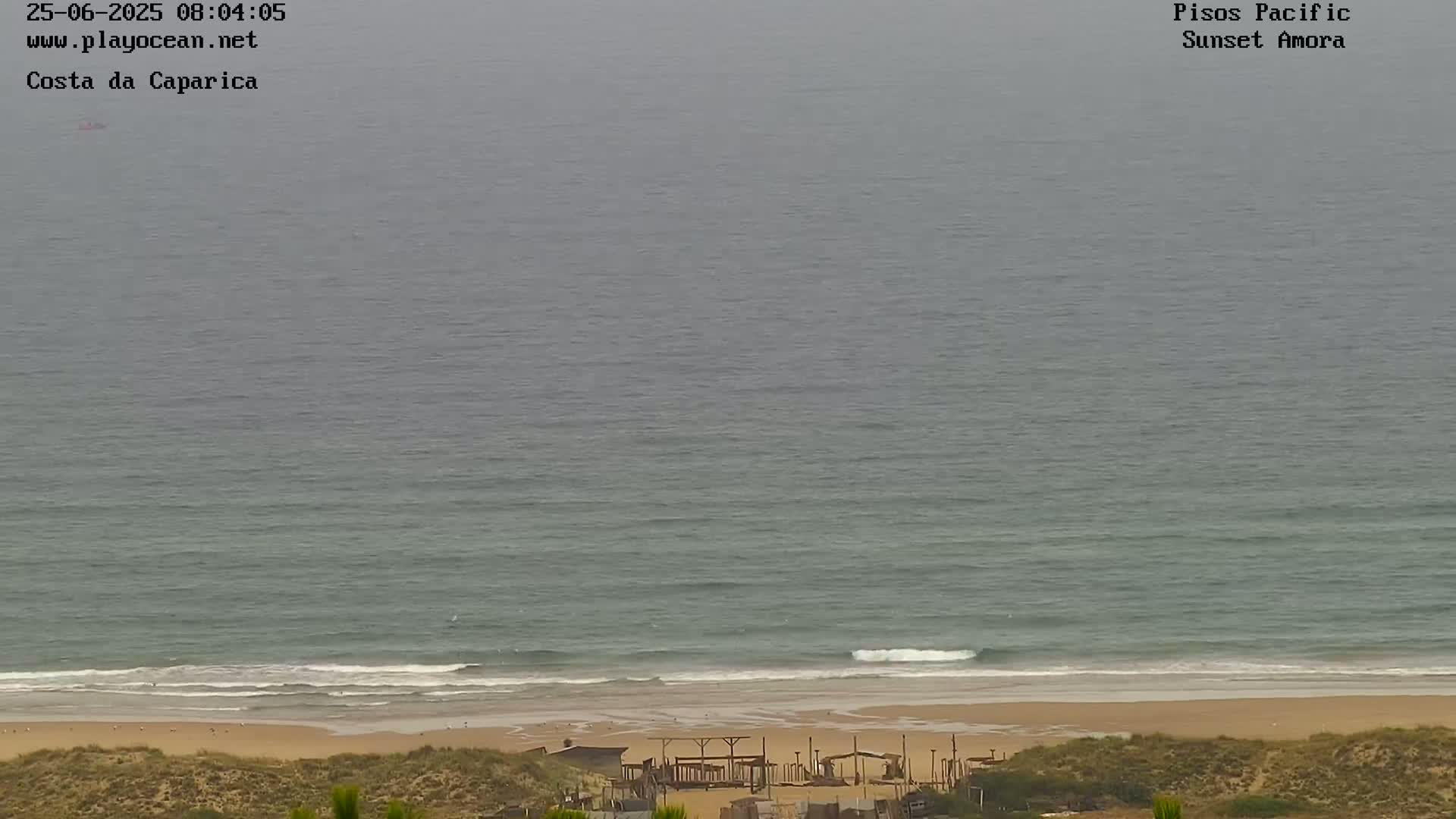 A mostly overcast sky overlooks a sandy beach with small waves rolling onto the shore and several structures visible near the shoreline.