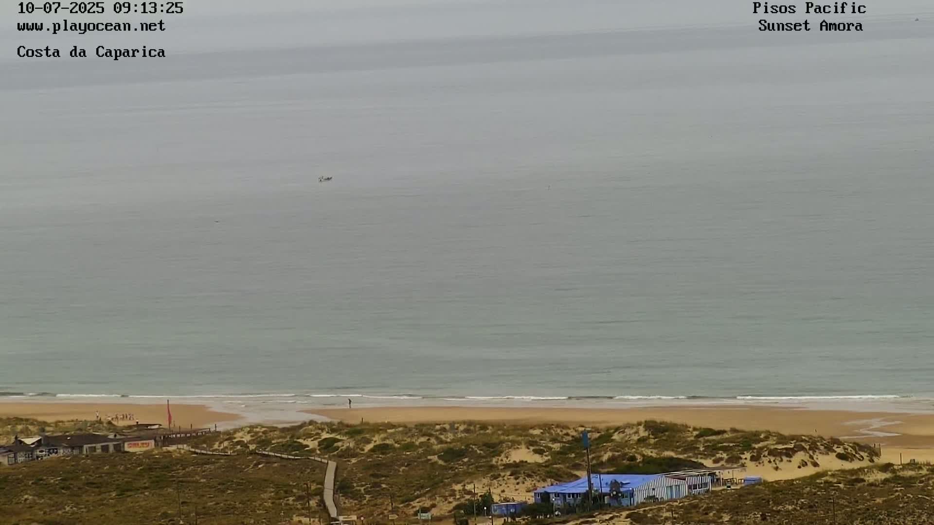 A calm, gray ocean meets a sandy beach backed by dune grasses and a small building, all under an overcast sky.