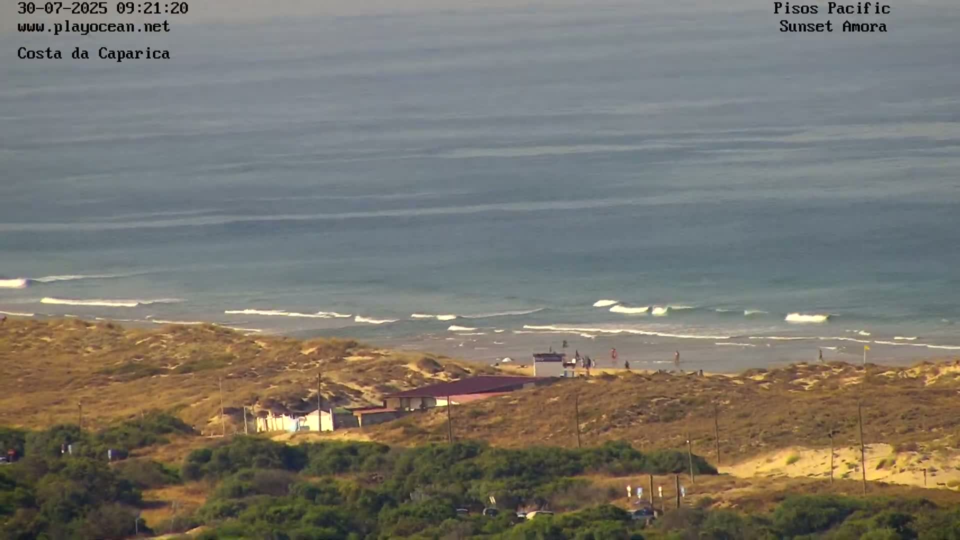 A sandy beach with a small building and people is visible in the foreground, with a calm ocean and small waves breaking in the background under a mostly clear sky.