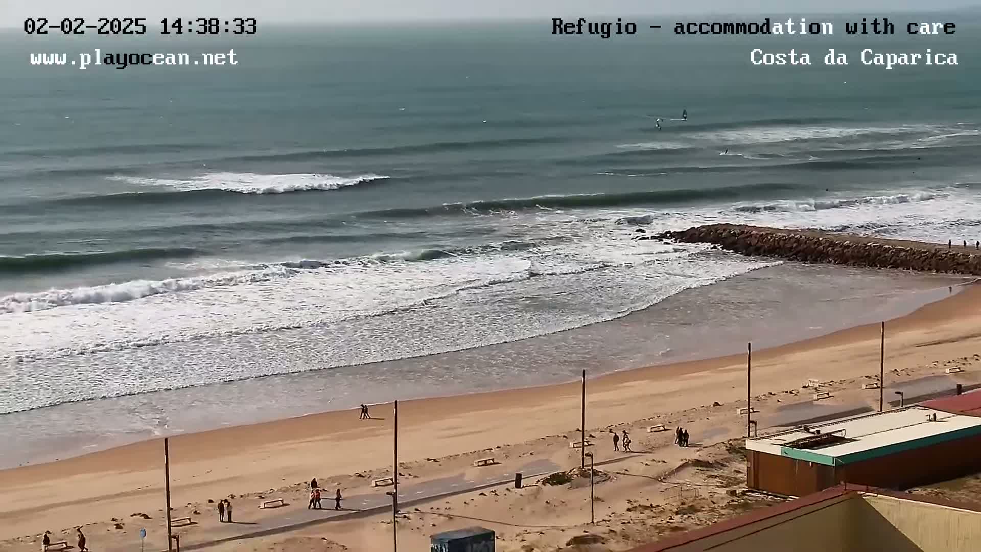A beach with several people walking along the shore and a few surfers in the ocean under an overcast sky.