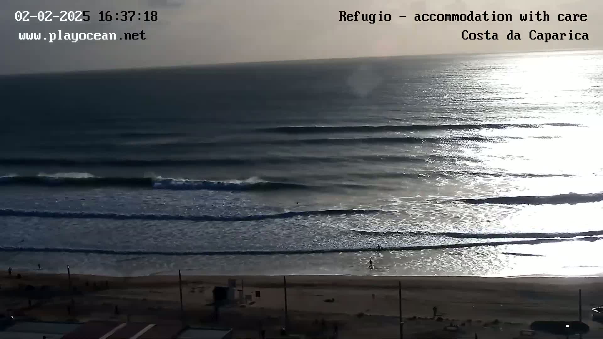 A long, sandy beach with several people on it and consistent waves breaking in the ocean under a partly cloudy sky.